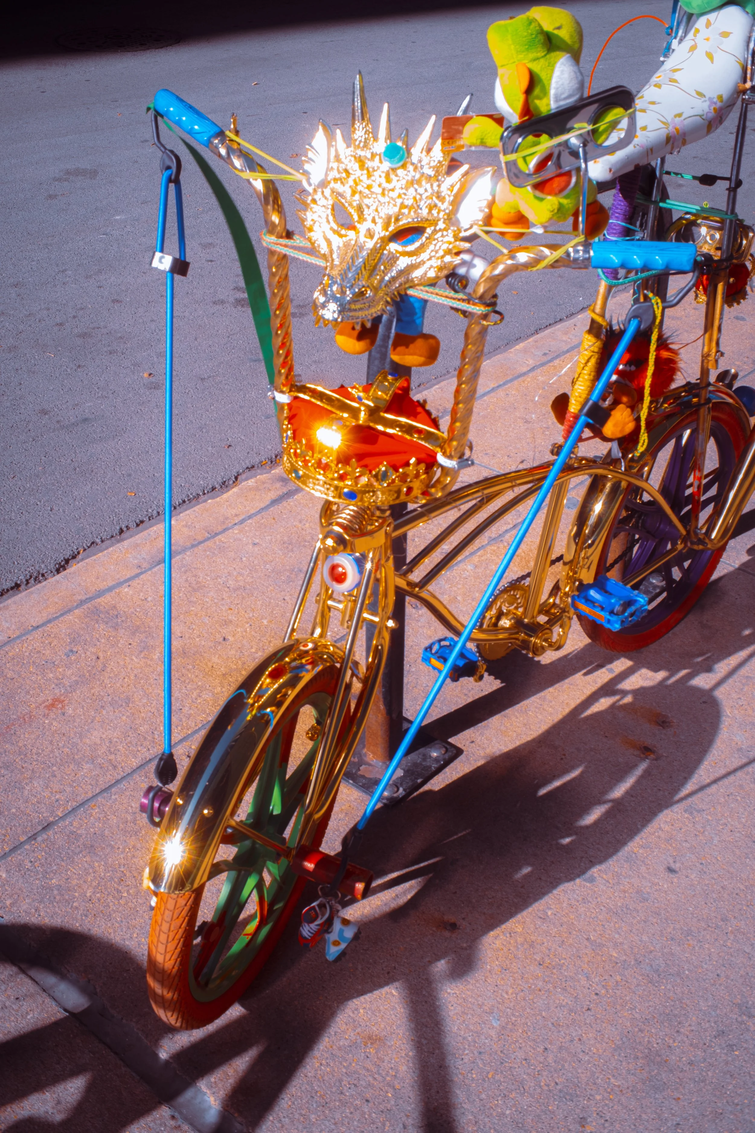 A bicycle decorated with a gold dragon face mask on the front, orange and green wheels, and stuffed toys attached with strings, parked on a sidewalk at dusk. The bike has colorful handle grips, and the toys include a green stuffed bear and a small pl