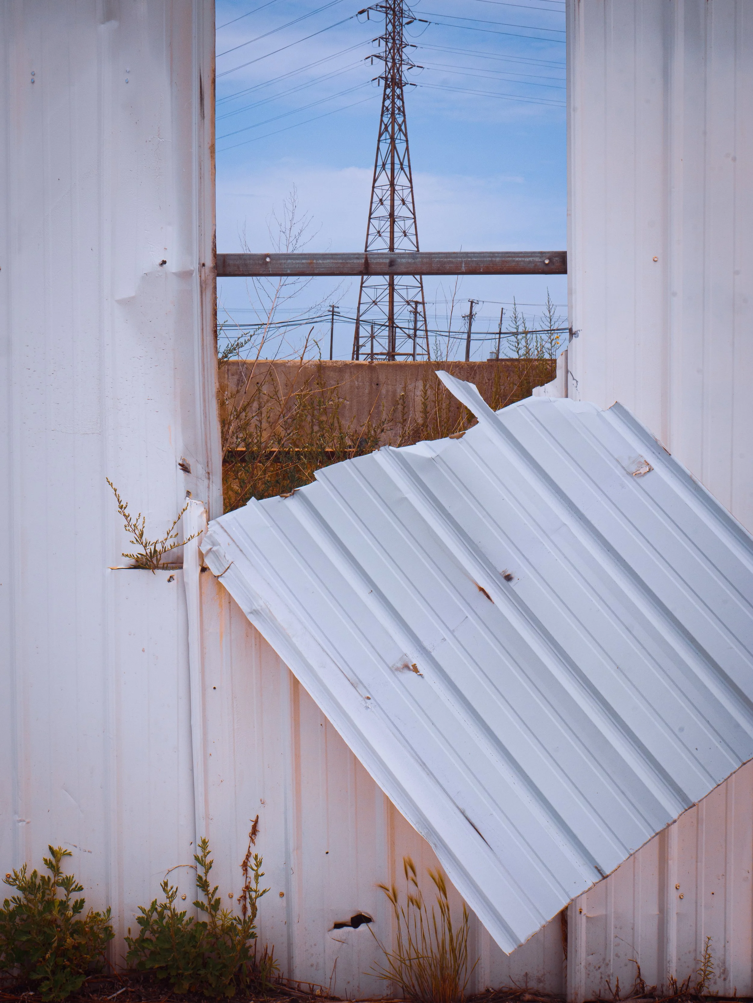 A white metal wall with a large, broken hole at the bottom left. A piece of corrugated metal leaning against the wall near a wooden beam. Behind the wall, a tall power transmission tower and power lines are visible against a partly cloudy sky.