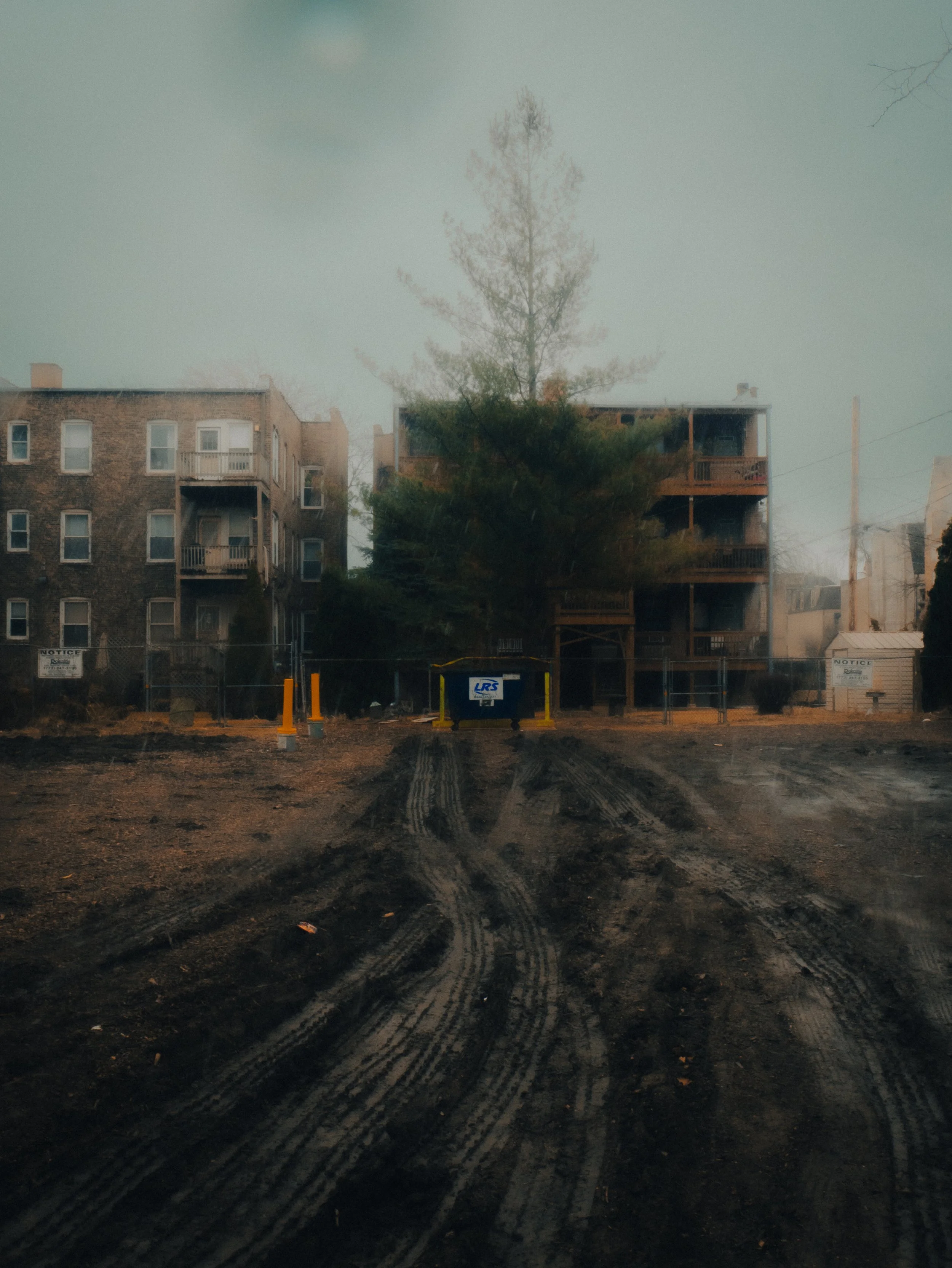 View of a muddy construction site road with tire tracks, a chain-link fence, multi-story brick apartment buildings, and a tree in front of the buildings, under a cloudy sky.