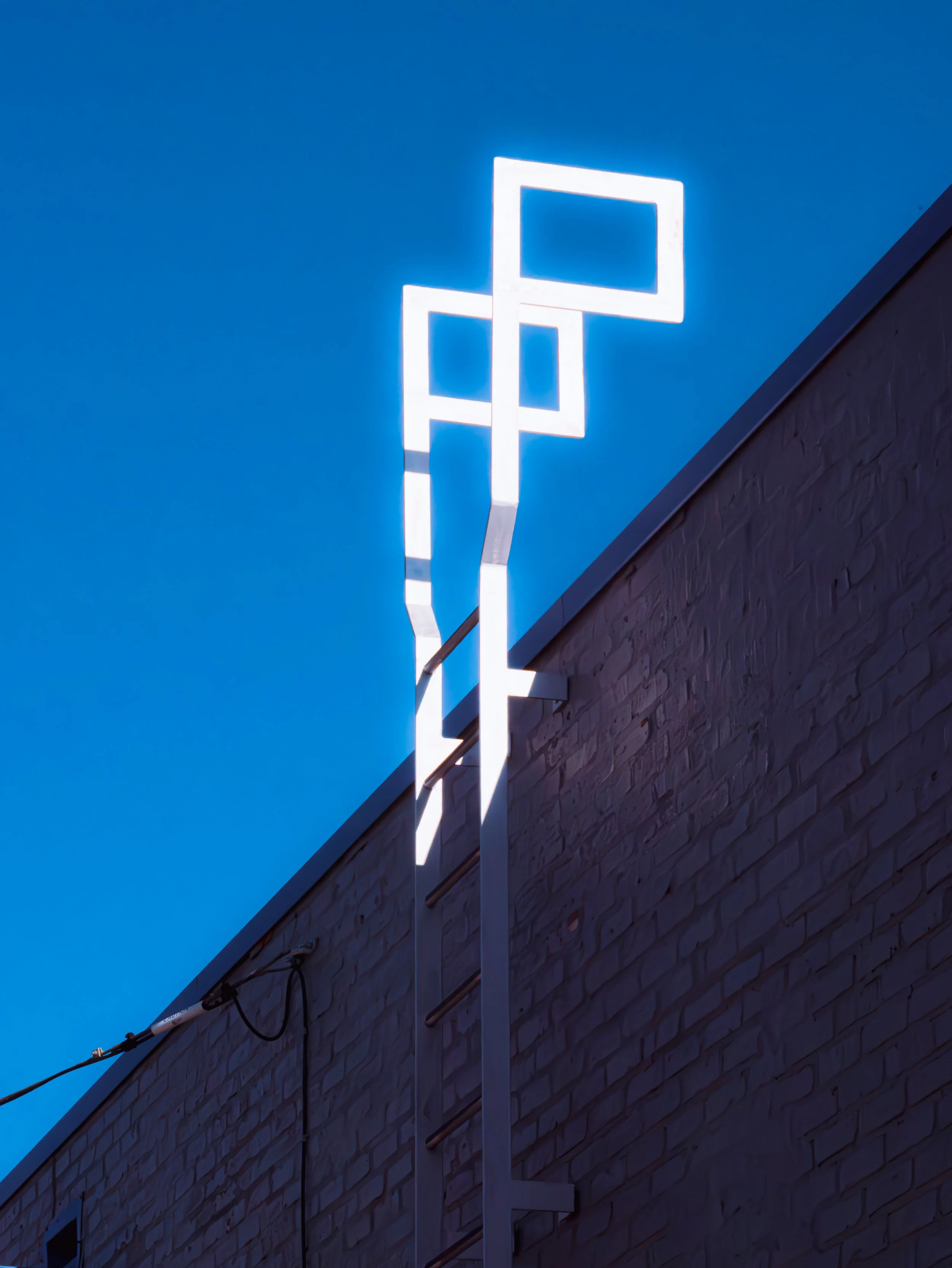 Outdoor wall with modern white neon art installation of overlapping squares against a blue sky background.