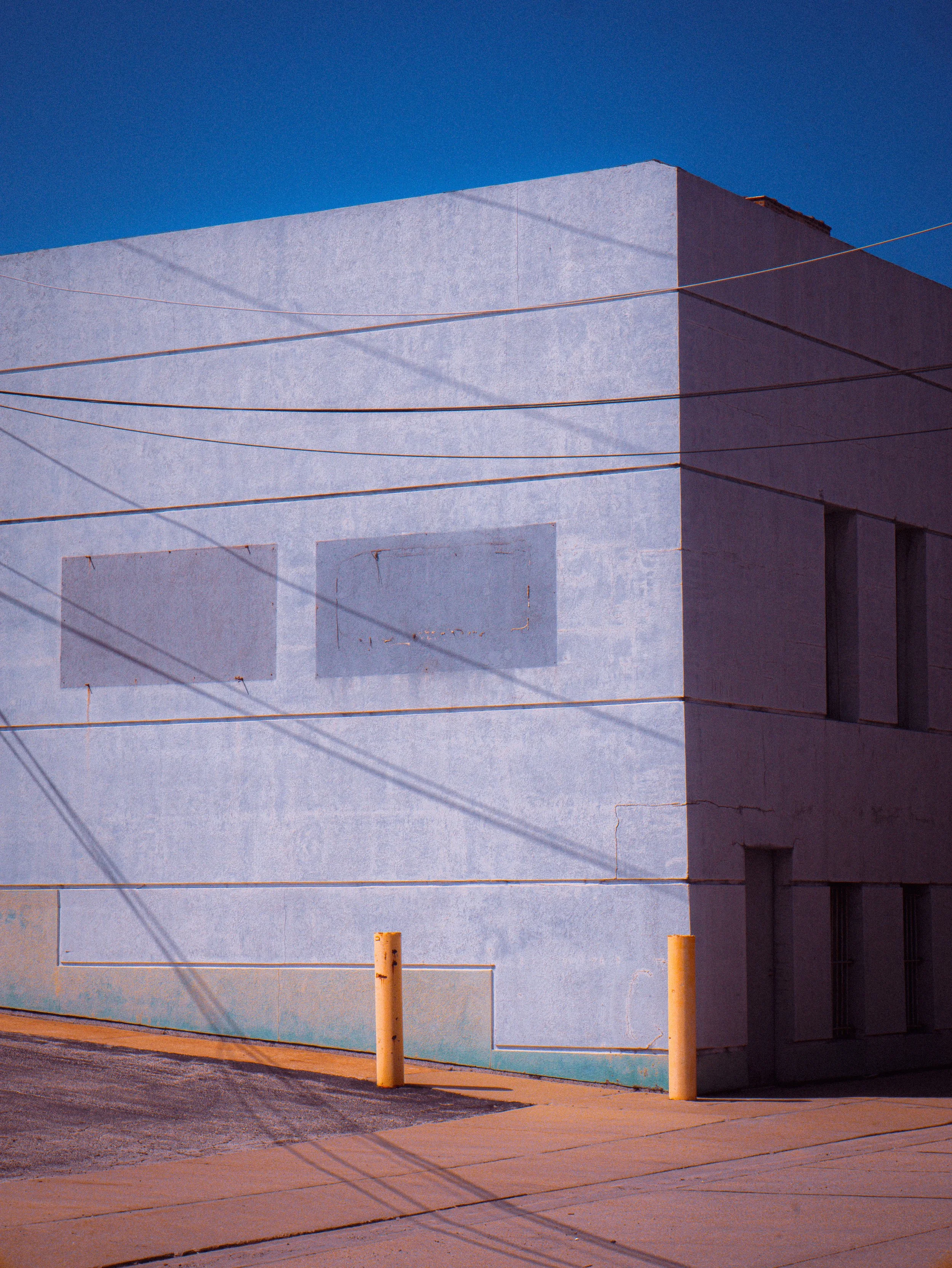 A white building with a textured exterior wall and small windows, set against a bright blue sky, with yellow bollards and visible power lines in the foreground.