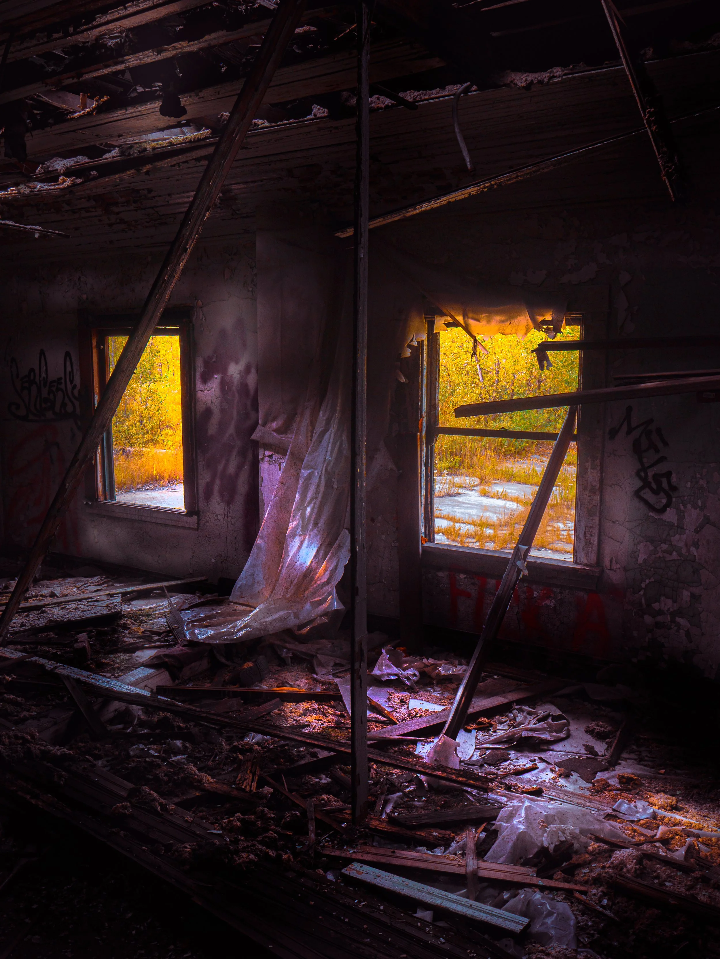 Interior of a destroyed building with collapsed roof and broken windows revealing trees and sunlight outside, debris and damage evident throughout.