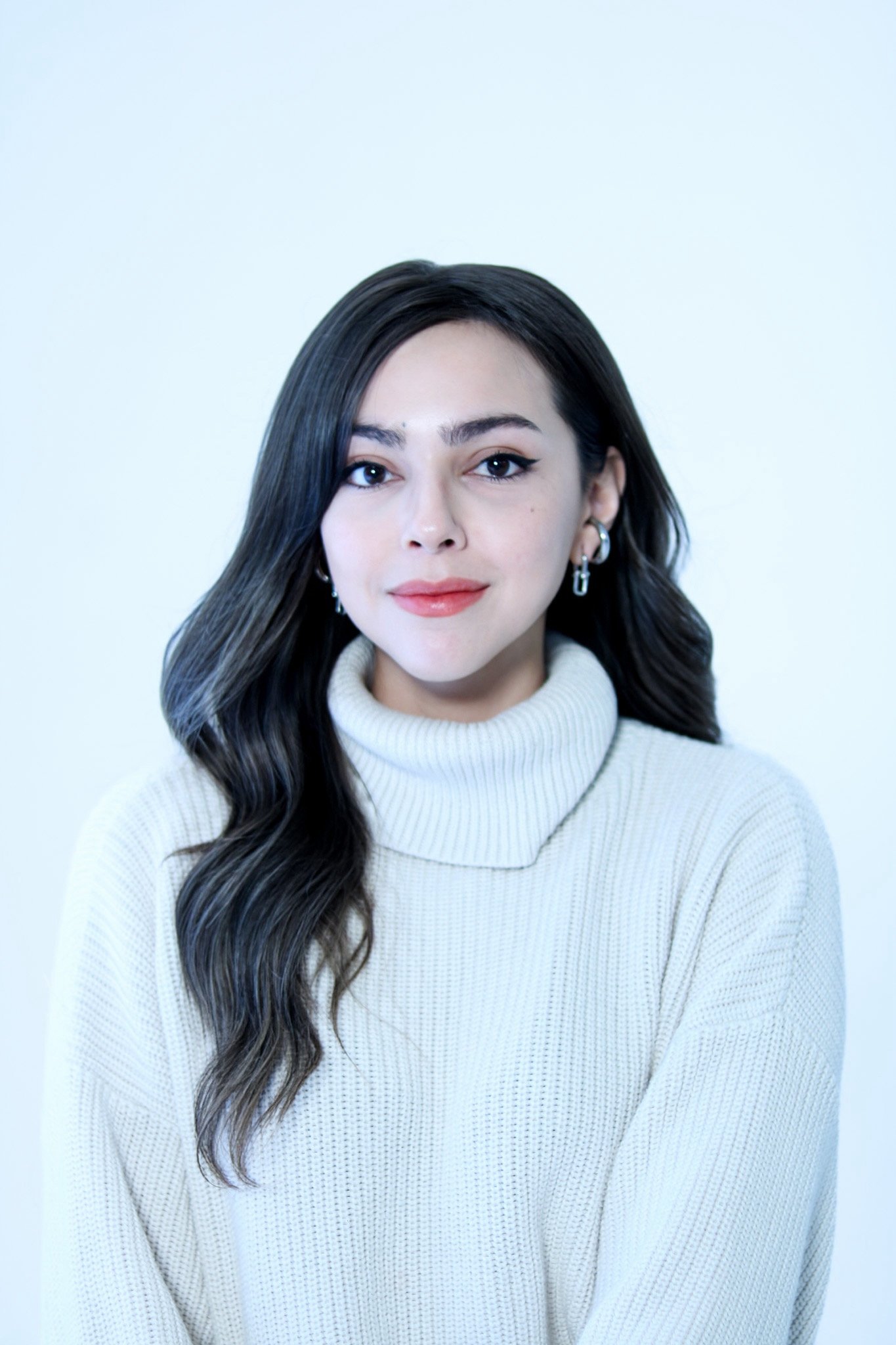 A young woman with long wavy dark hair, wearing a cream-colored turtleneck sweater and earrings, looking directly at the camera with a neutral expression and a slight smile, against a plain light blue background.
