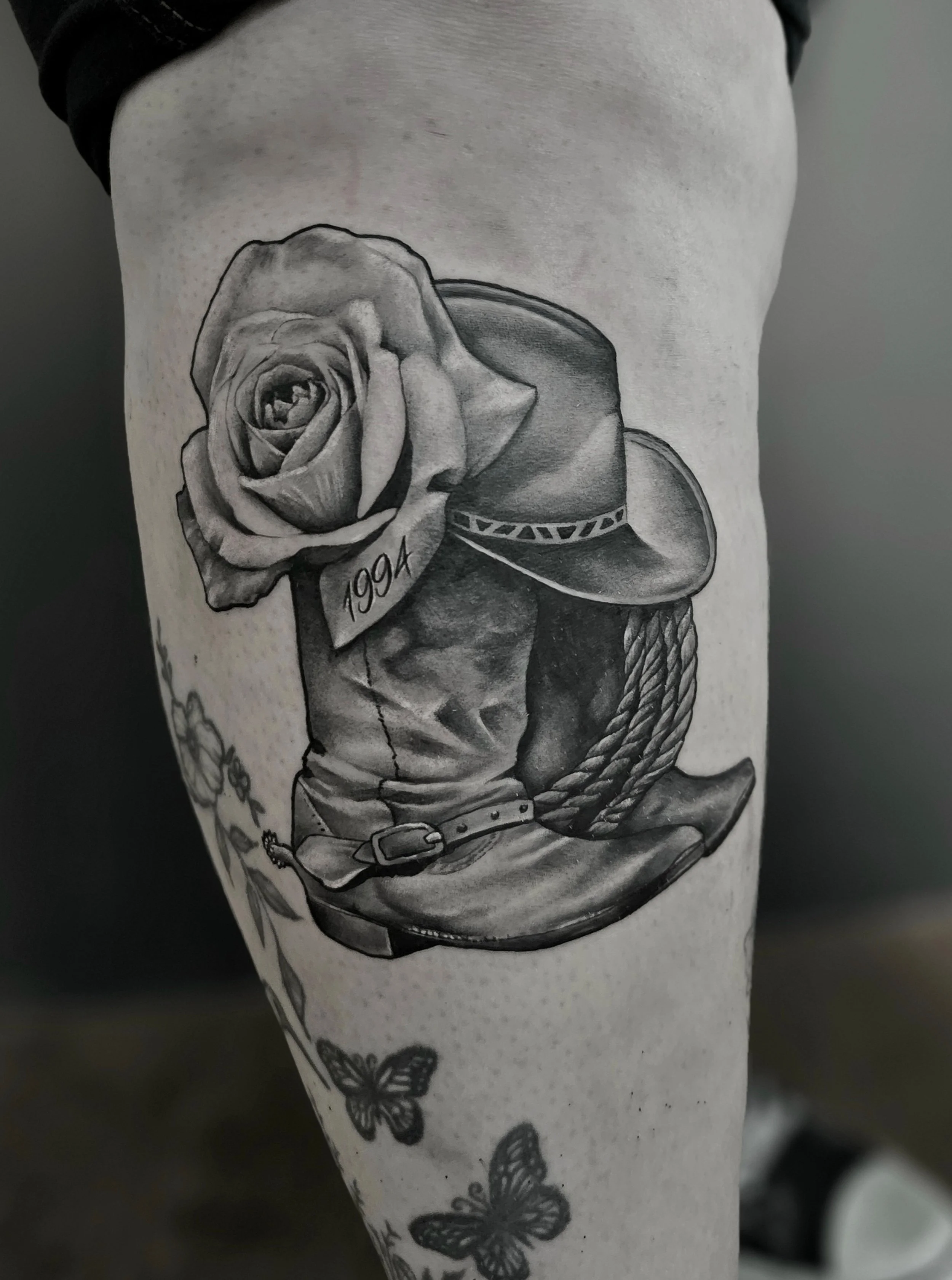 Black and grey tattoo featuring a detailed rose with the year '1994' beneath it, a cowboy hat with a patterned band, and a coiled rope, with additional butterflies and floral elements.
