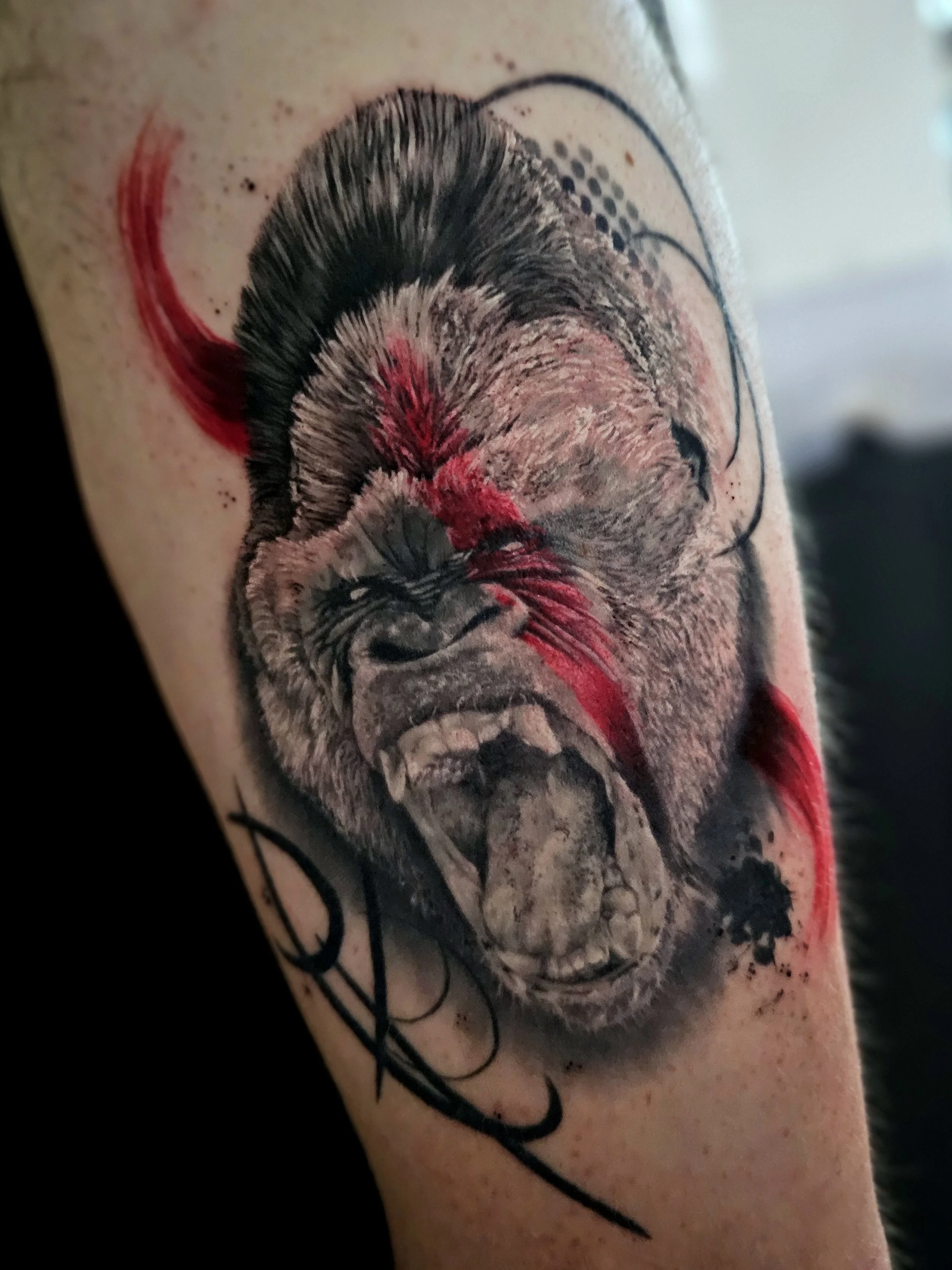 A tattoo of a realistic lion's face on a person's arm, with detailed fur, eyes, and nose, and red and black ink accents.
