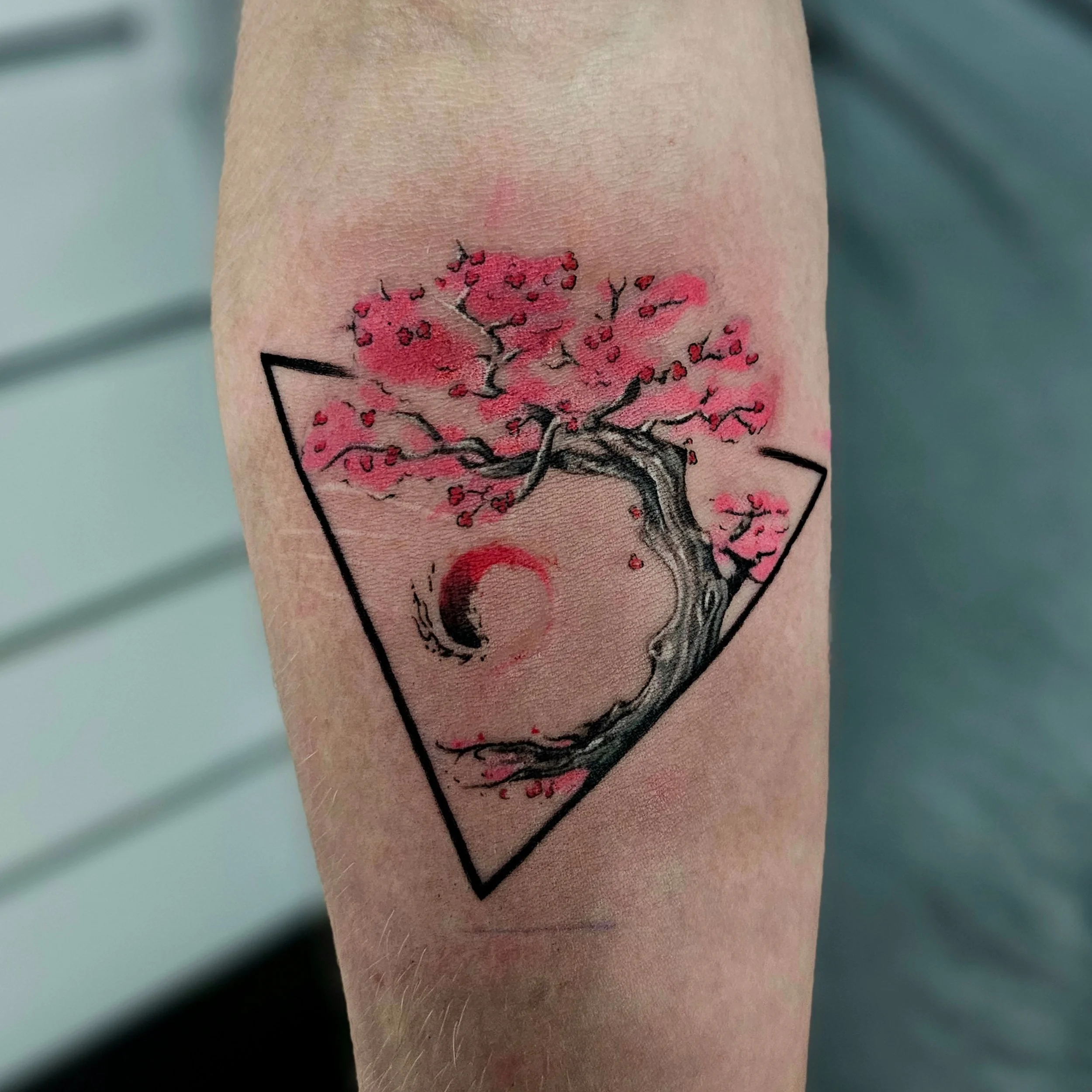 Close-up view of a tattoo on a person's arm, depicting a tree with pink blossoms and black branches within a black geometric border.