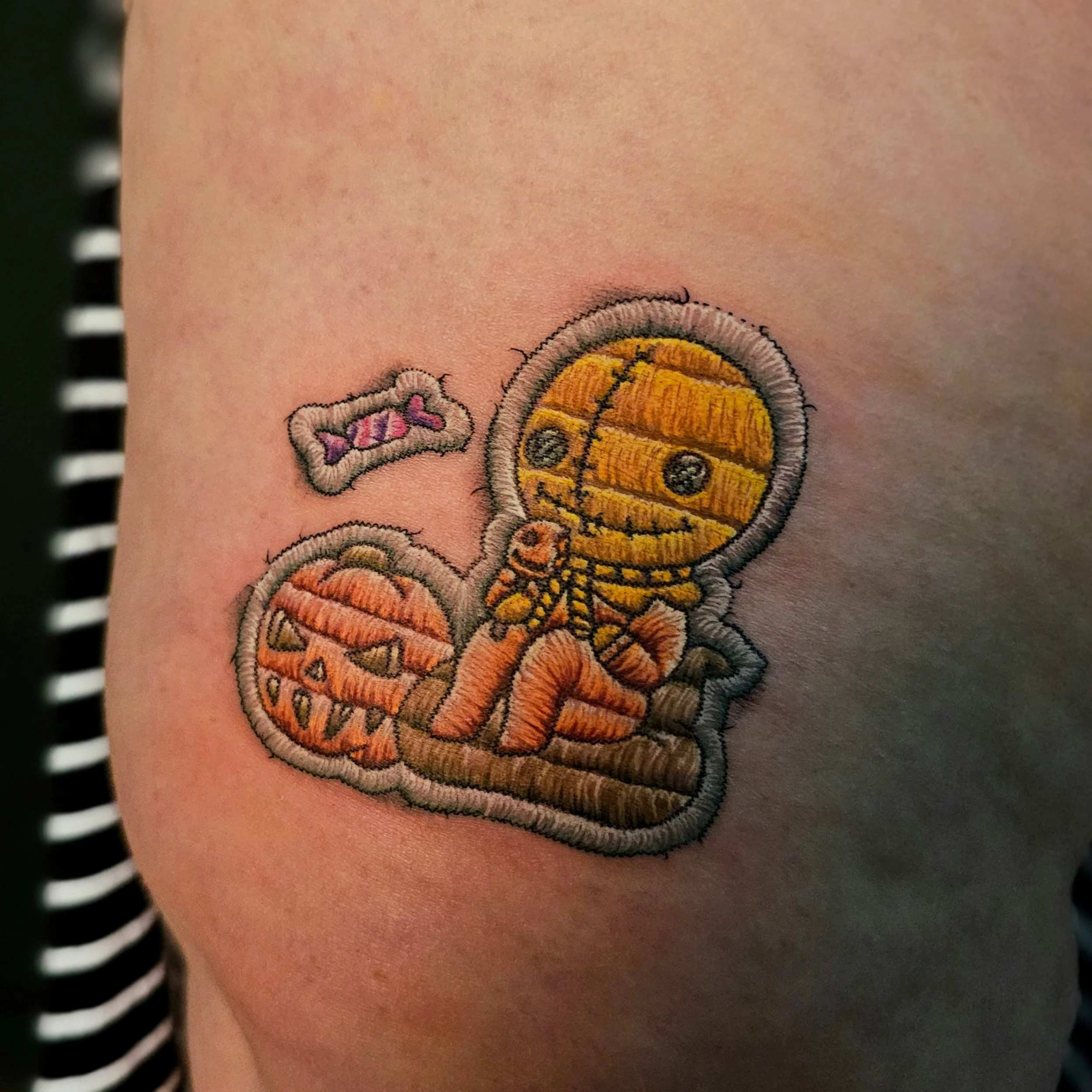 Close-up of a colorful embroidered flower tattoo on skin, with part of a black and white striped garment visible on the left side.