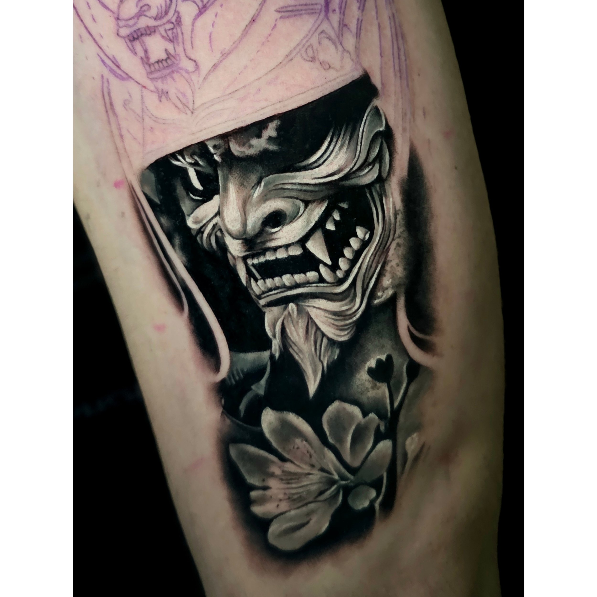 Close-up of a tattoo featuring a black and white smoking skull and a partially visible face, with tattoo sketches on skin in the background.
