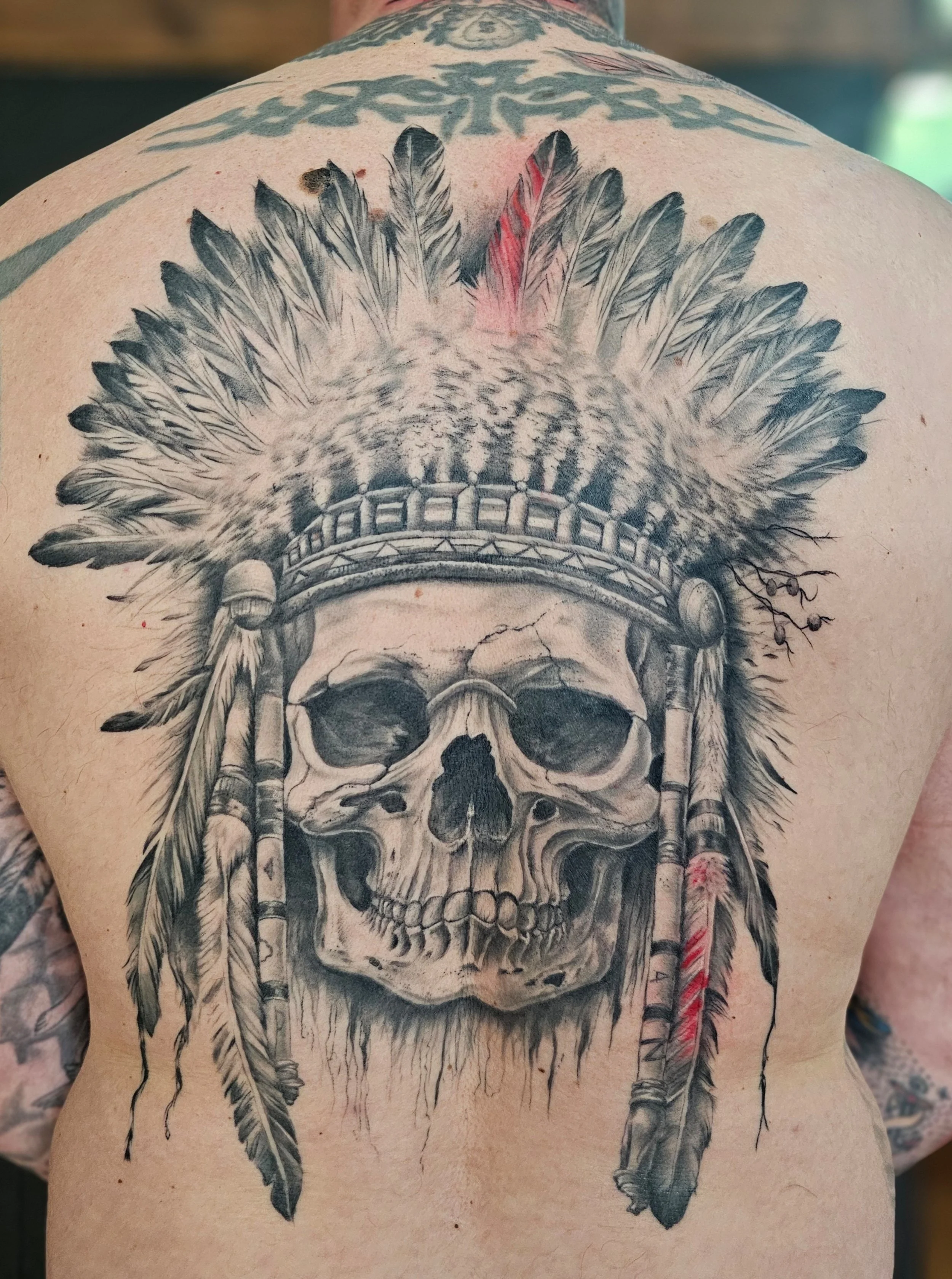 A person's back with a detailed black and gray tattoo of a human skull adorned with a feather headdress and feathers hanging down.