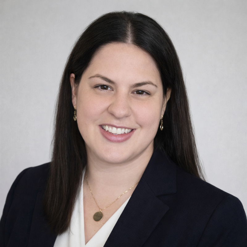 Professional headshot of a woman with long dark hair, smiling, wearing a navy blazer, white top, and gold jewelry.