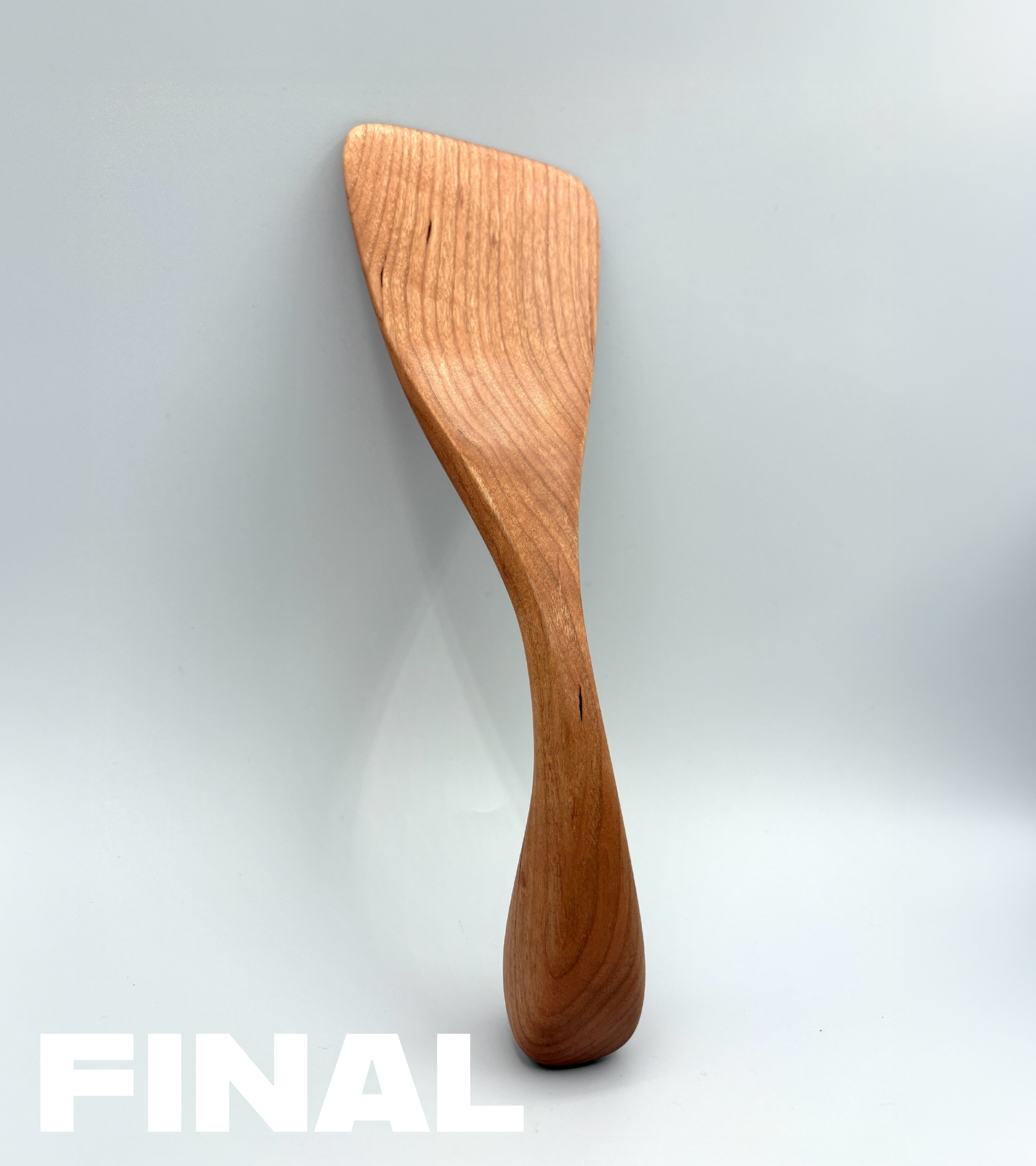 Close-up of a wooden spatula with a rounded head and curved handle against a light background, with the word 'FINAL' partially visible at the bottom.