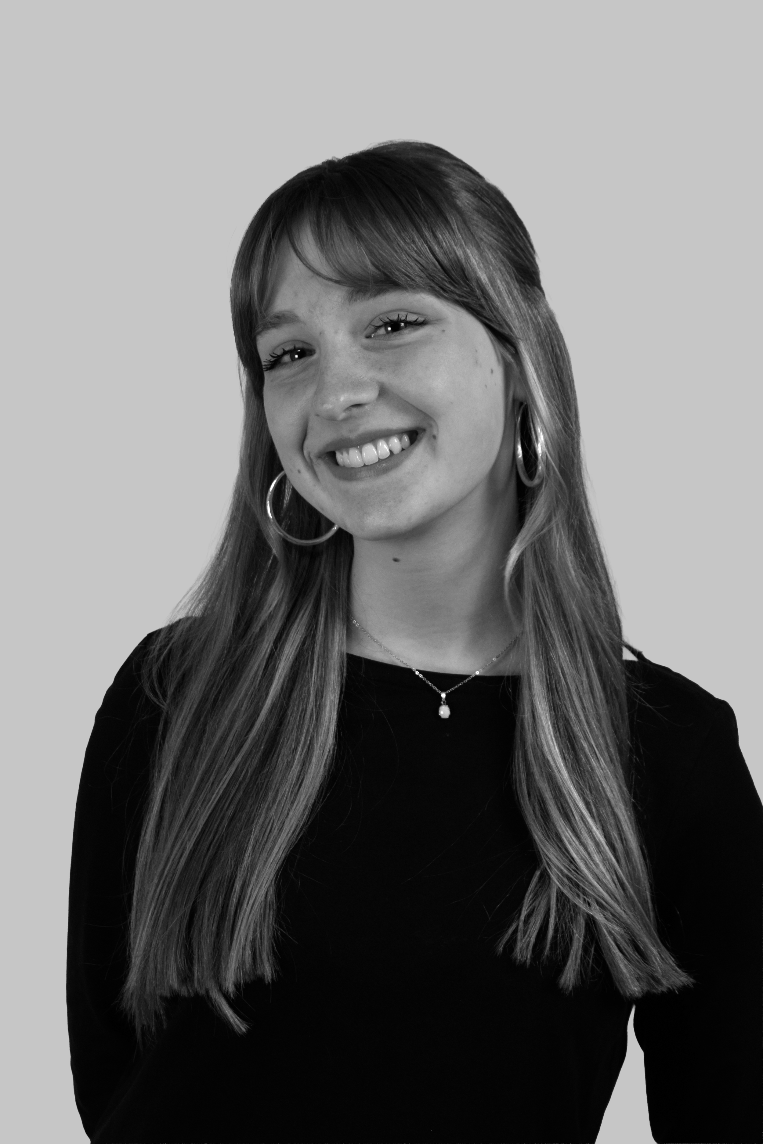 Black and white portrait of a smiling young woman with long hair, wearing hoop earrings and a necklace, against a plain backdrop.