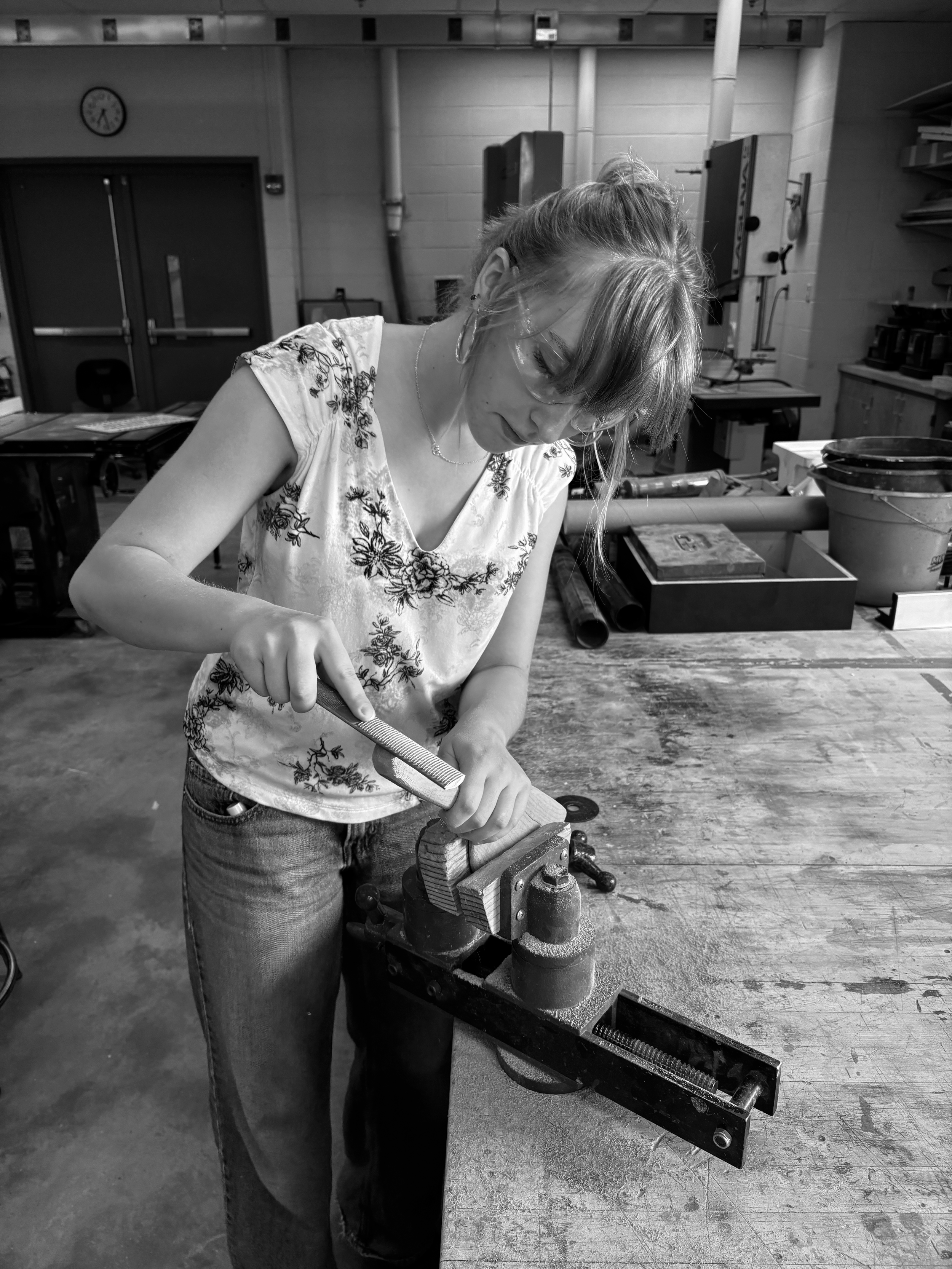 A woman in a workshop using a hand file on a metal piece clamped in a vise, surrounded by tools and equipment.