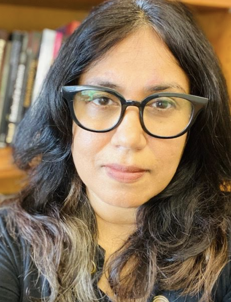 Close-up portrait of a woman with long dark hair and black eyeglasses in front of a bookshelf