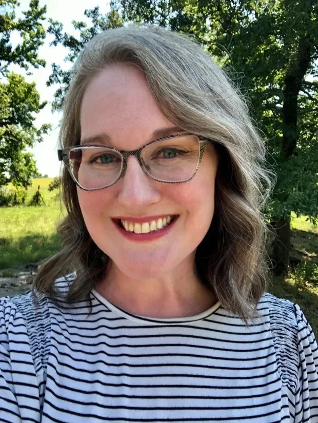 Smiling woman wearing glasses and a striped shirt outdoors in a sunny wooded setting