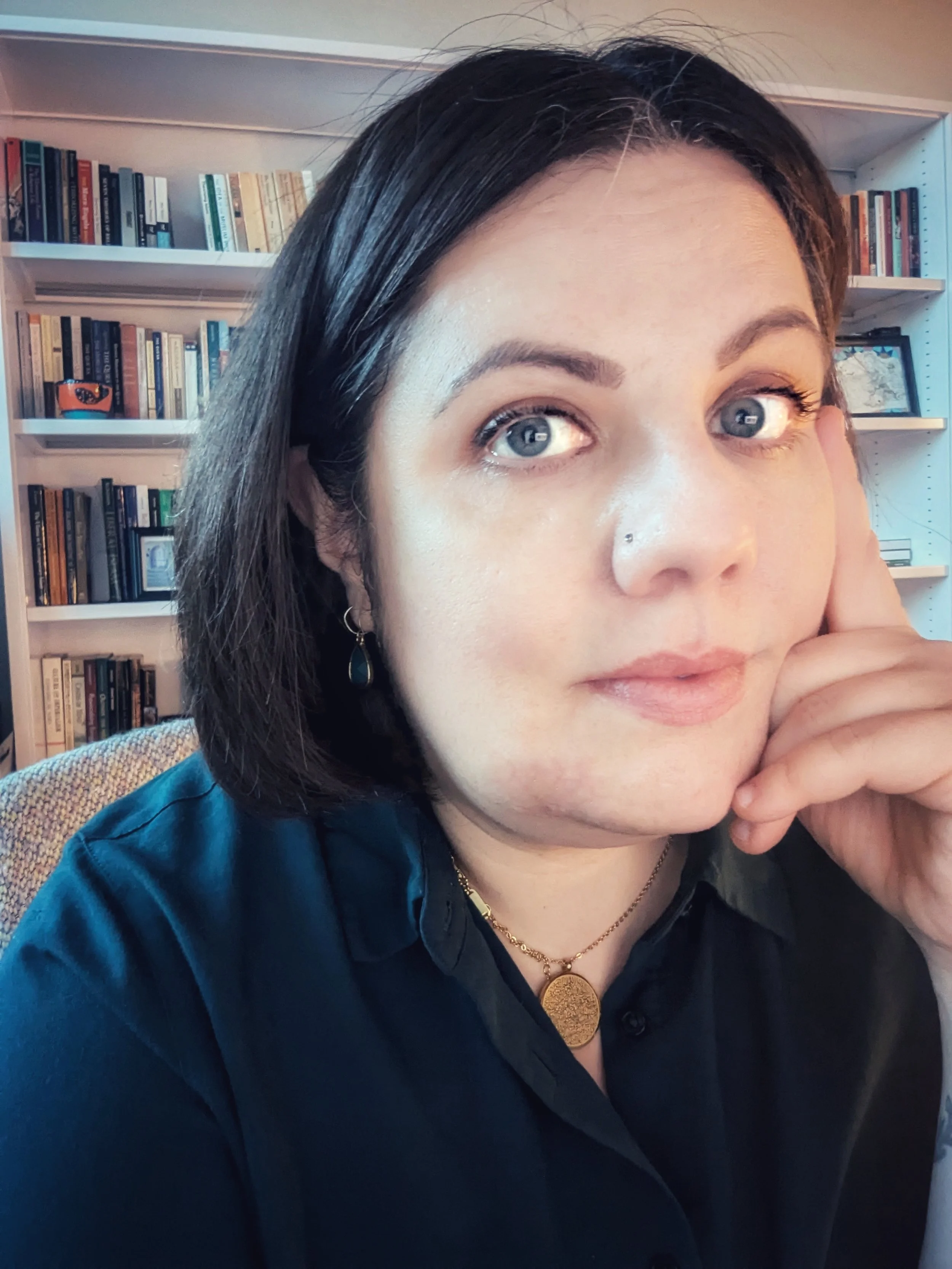 Close-up portrait of a woman with dark hair, blue eyes, jewelry, and a black blouse sitting in front of bookshelves.