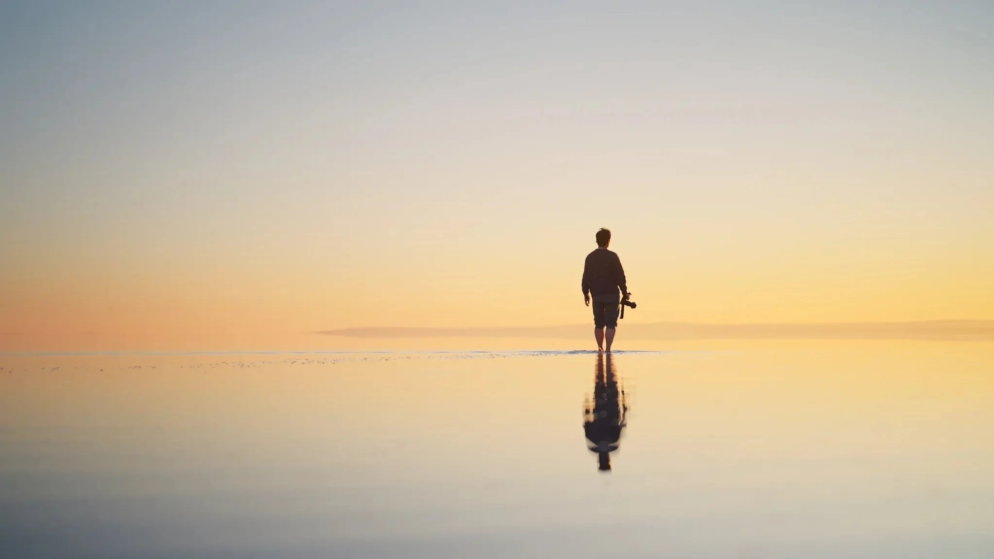 A person walking across a sunlit beach, reflected in the shallow water