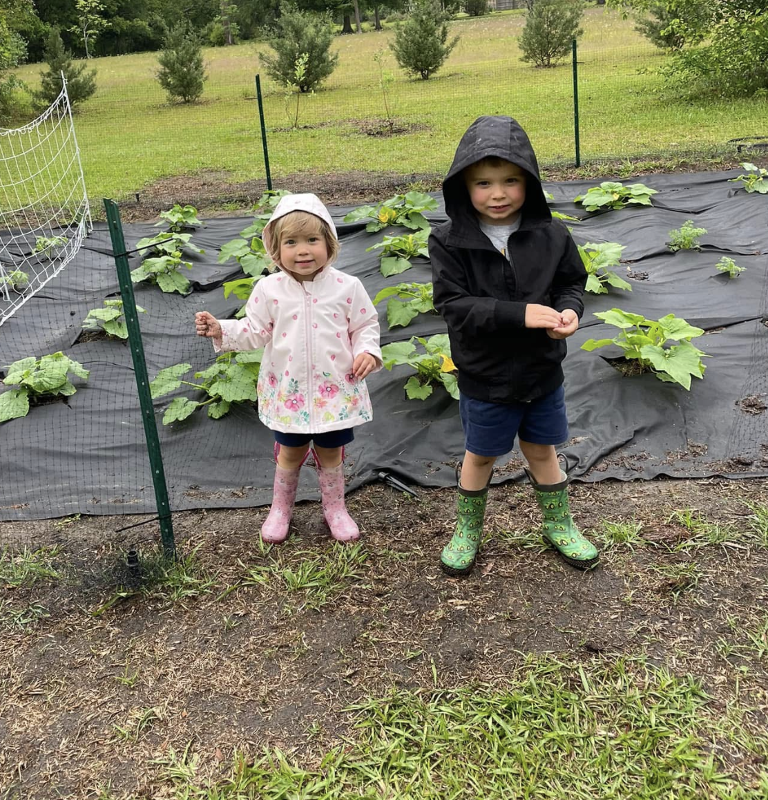 Two toddlers in rain boots in a backyard vegetable garden