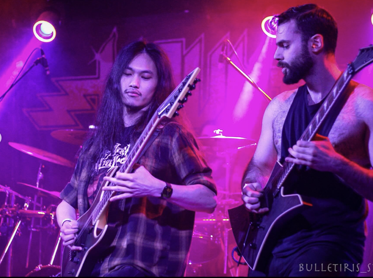 Two male musicians playing electric guitars on stage with purple lighting and a drum set in the background.