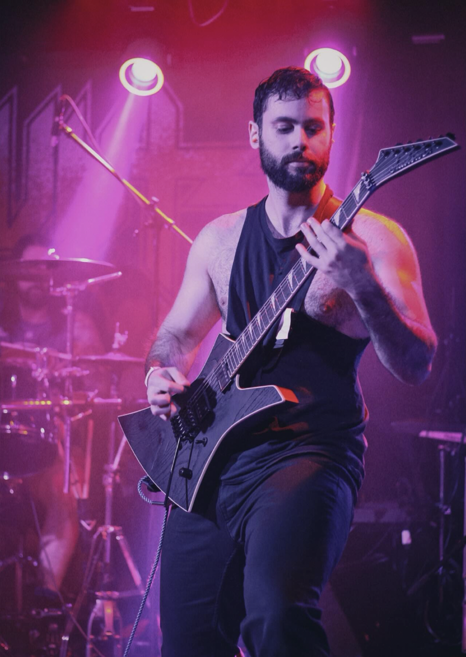 A man playing an electric guitar on stage with purple and pink lighting, wearing a black sleeveless shirt, in front of a drum set.
