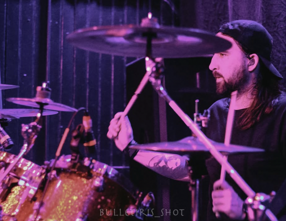 A man with long hair, a beard, and a black cap playing drums in a dimly lit, purple-toned venue.