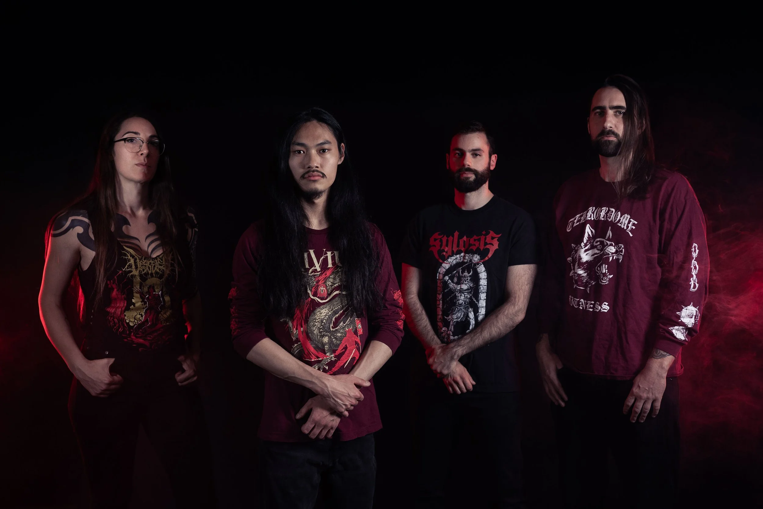Group portrait of four men and women with long hair and beards, standing against a black background with red lighting, dressed in black and dark red band shirts with graphic designs.