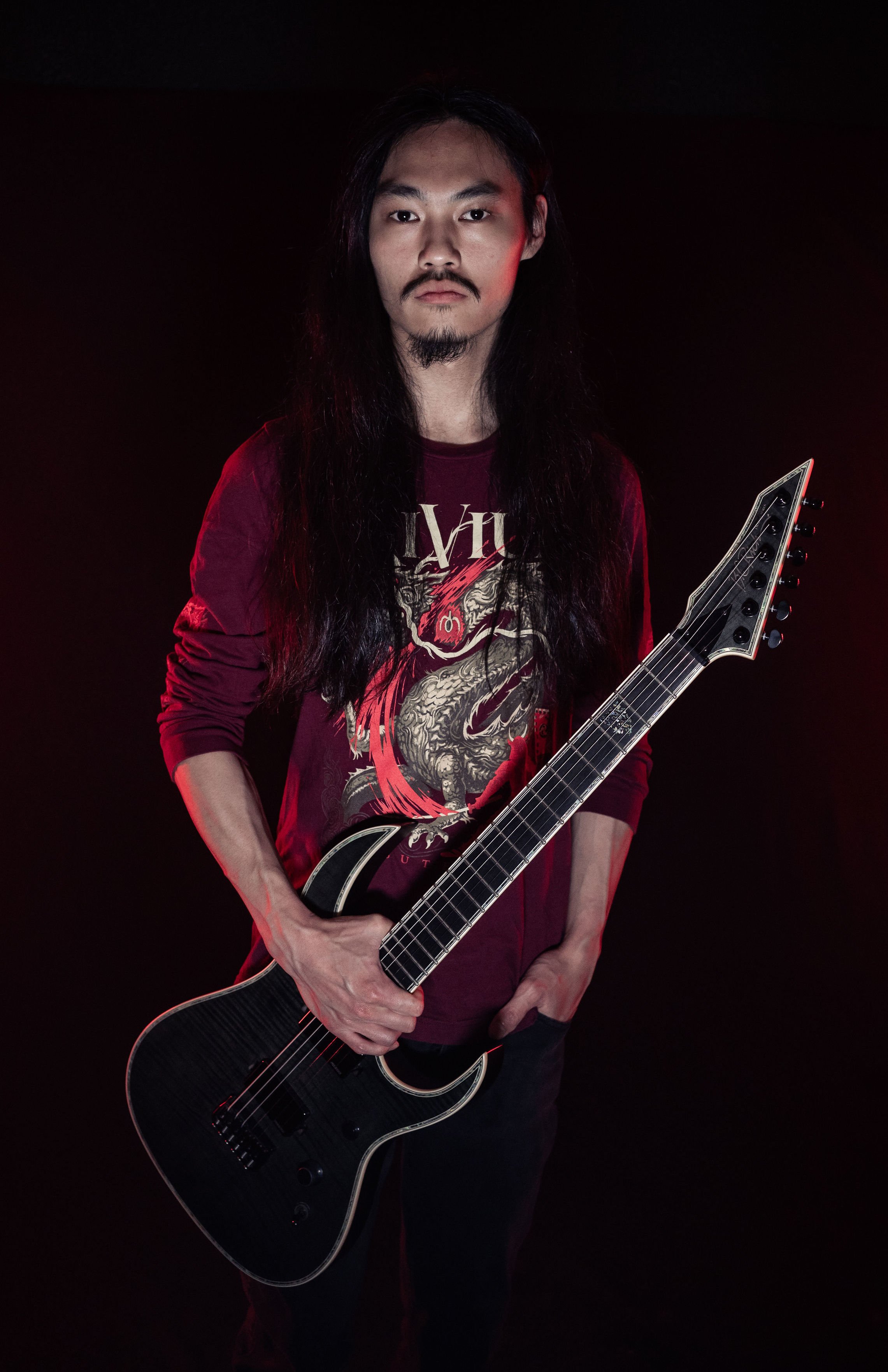 A man with long black hair, a mustache, and a goatee holding a black electric guitar, wearing a red graphic long-sleeve shirt, standing against a dark background with red lighting.