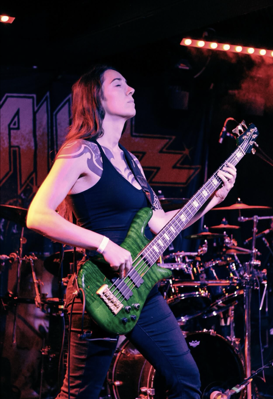 A woman playing a green electric bass guitar on stage with a drum set behind her, under colorful stage lighting, in a rock concert setting.