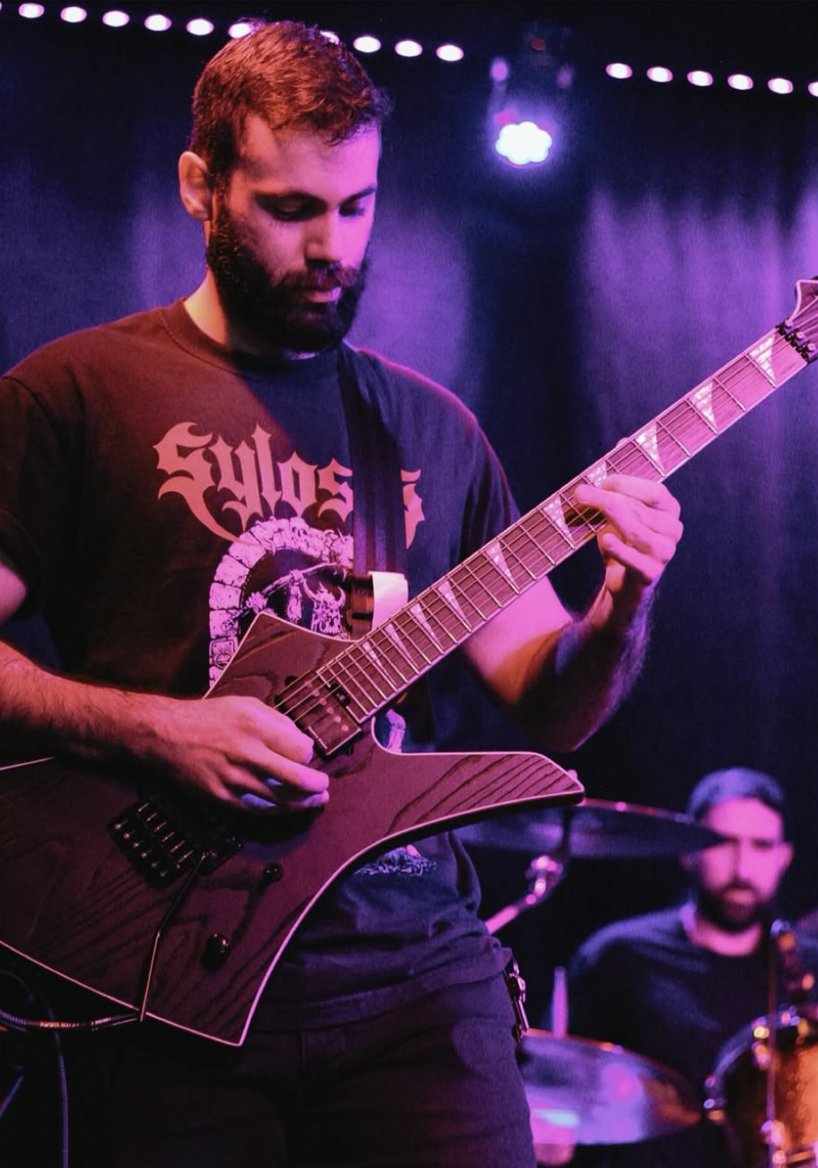 A bearded man with short hair playing an electric guitar on stage with purple and blue lighting. In the background, a drummer is visible.