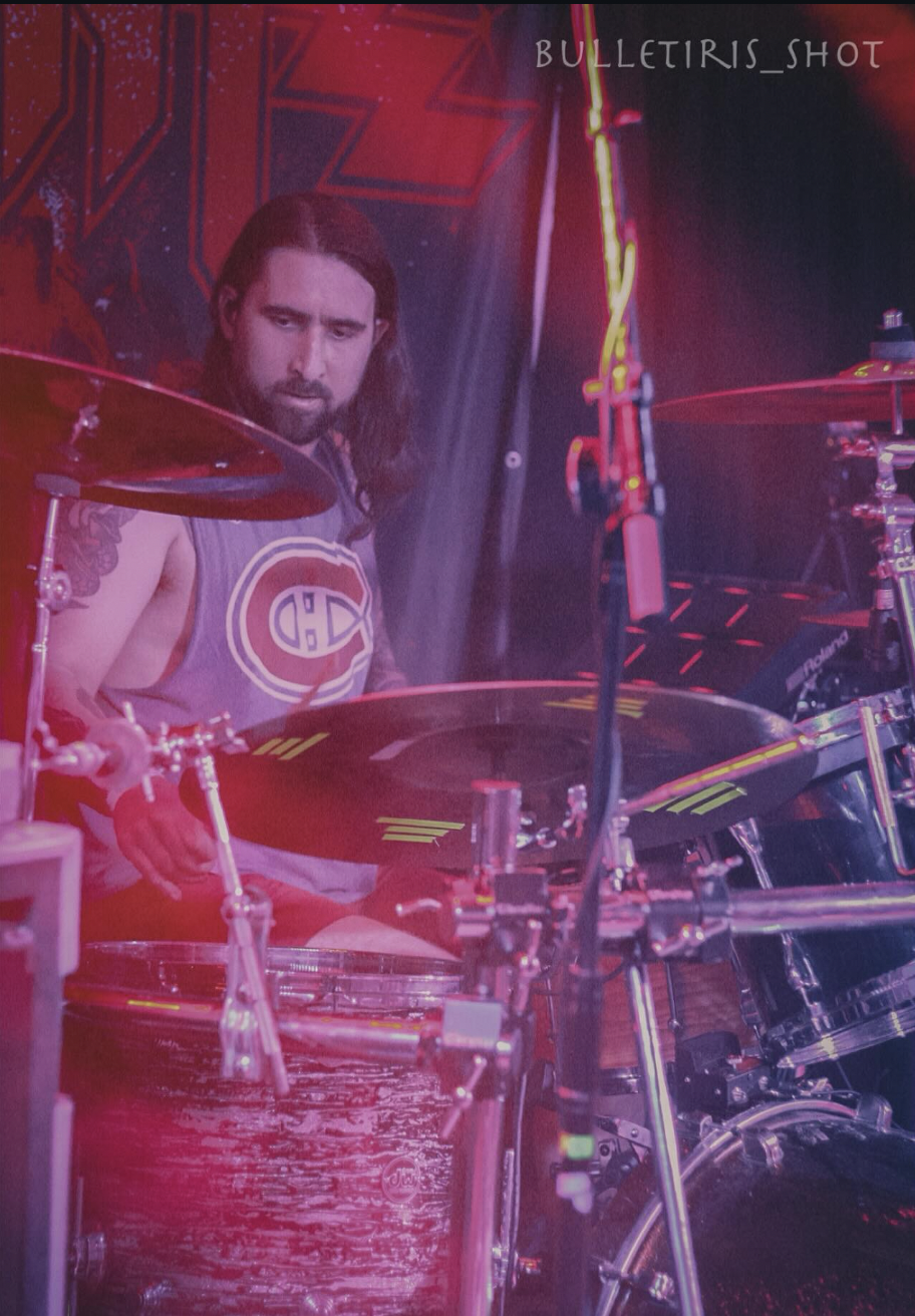 A man with long hair and a beard playing a drum set on stage, wearing a sleeveless shirt with a sports logo, surrounded by stage lights and musical equipment.