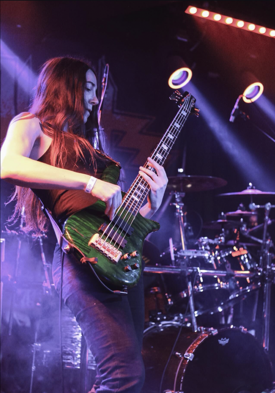 A young woman playing an electric bass guitar on stage with colorful lights and a drum set in the background.