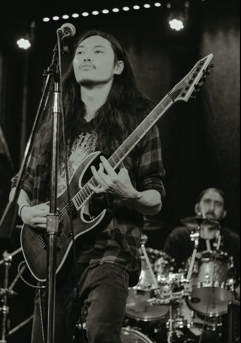 A black and white photo of a long-haired male musician playing an electric guitar and singing into a microphone on stage, with a drummer visible in the background.