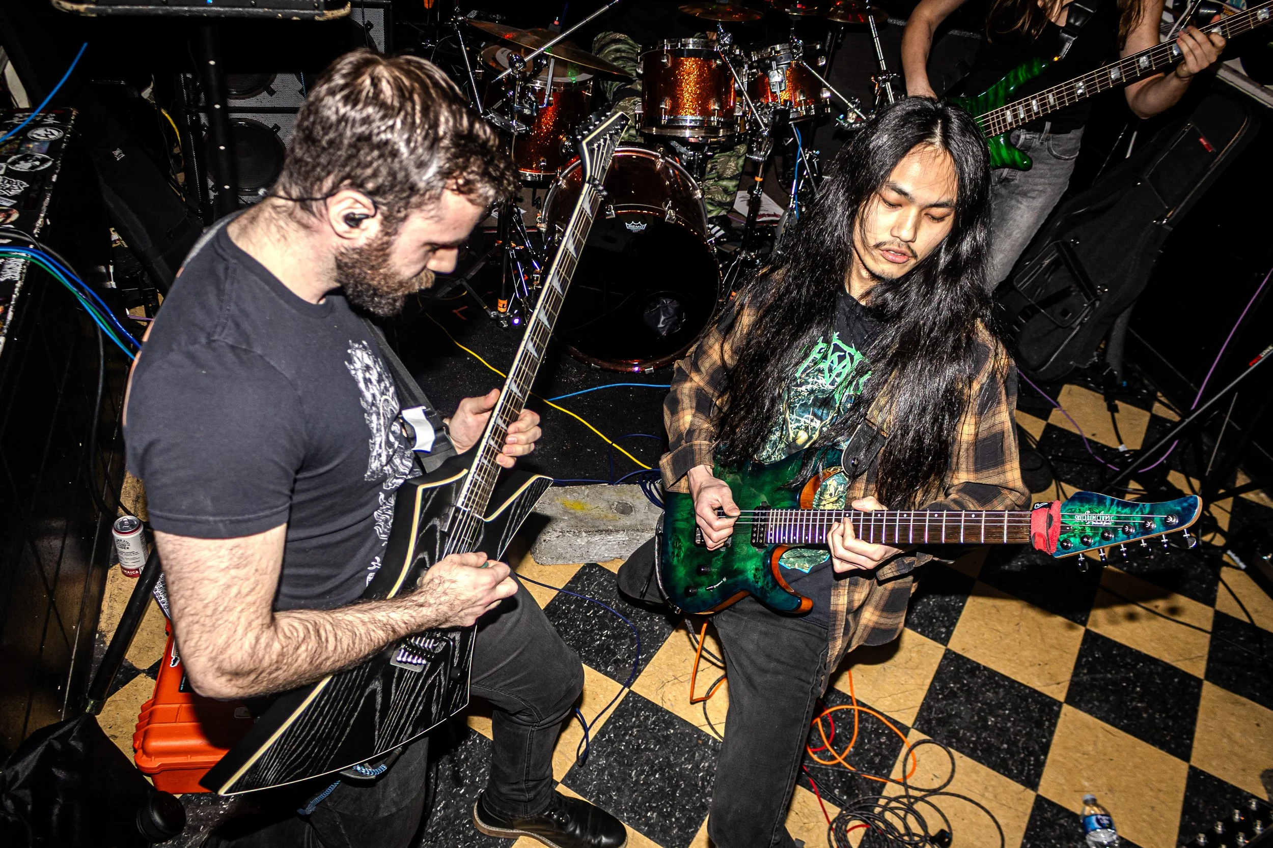 Two musicians playing electric guitars and a drummer in the background during a live performance.