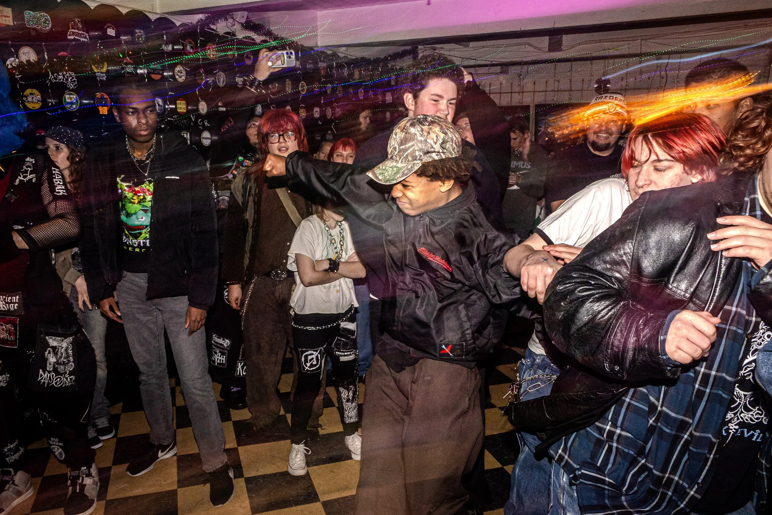 Group of young people, some with colorful hair and tattoos, dancing and socializing in a dimly lit room decorated with string lights and stickers on the wall. The scene appears lively with movement and energy.