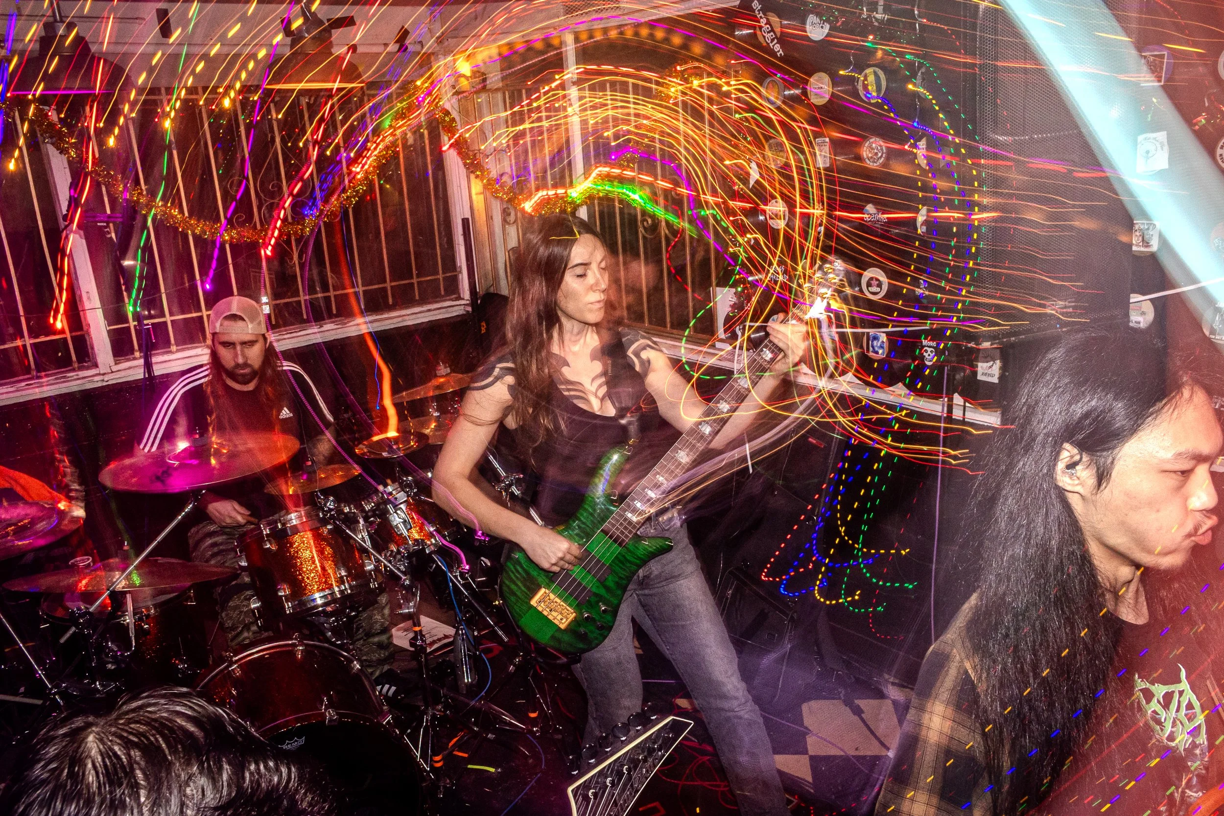 A woman playing an electric guitar and a man on drums performing at a lively, colorful party with light trails and decorations in the background.