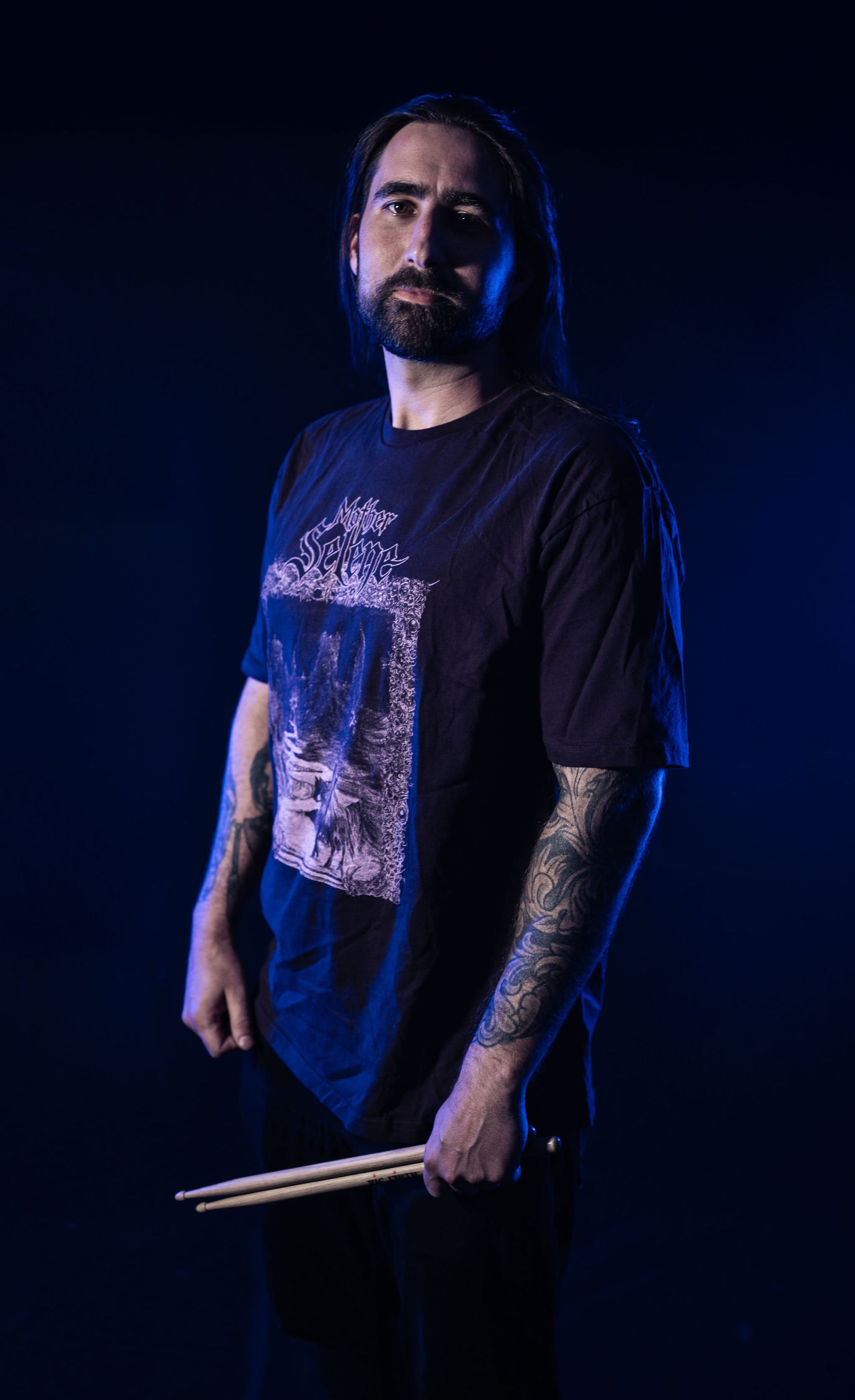 A man with long dark hair, a beard, and visible tattoos on his arms holding drumsticks, wearing a black t-shirt with a graphic design, standing against a dark blue background.