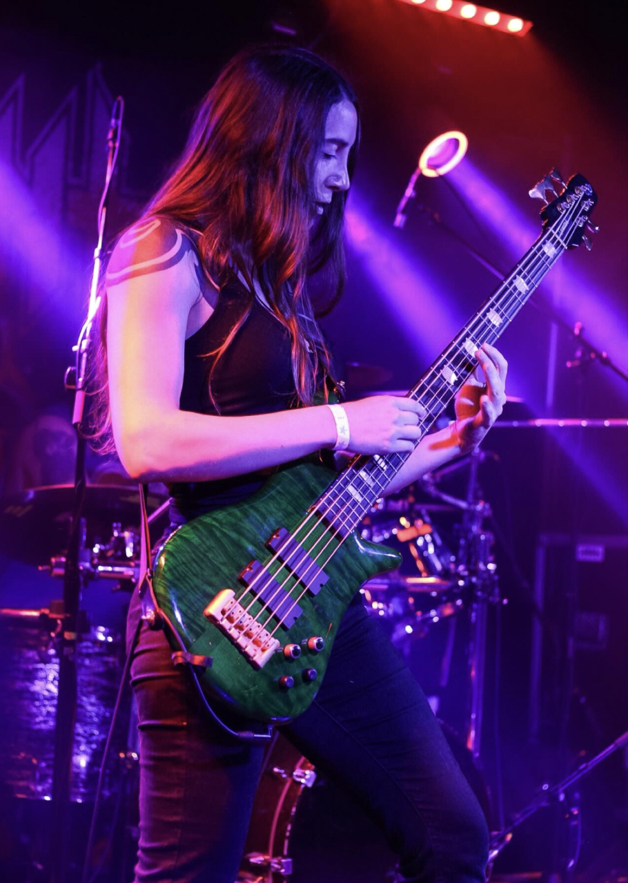 A woman playing a green electric bass guitar on stage under colorful purple and pink lights.
