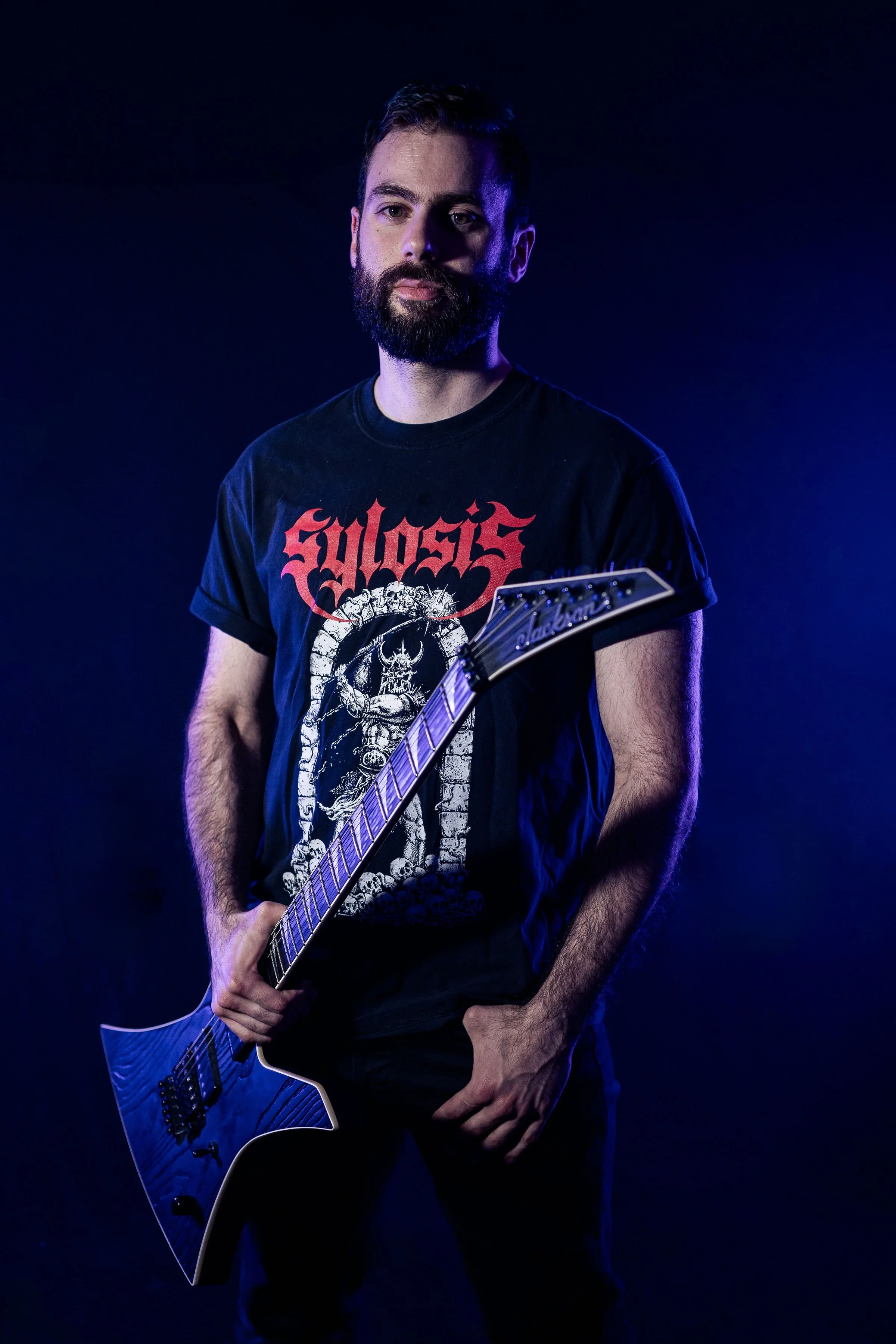 A man with a beard wearing a black t-shirt with red and white graphics, holding a black electric guitar against a dark background with blue lighting.