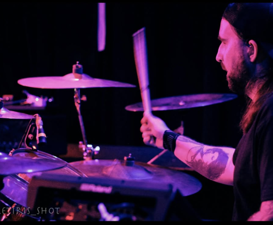 A musician playing drums on stage with purple lighting, focusing on the drummer's profile and tattoos.
