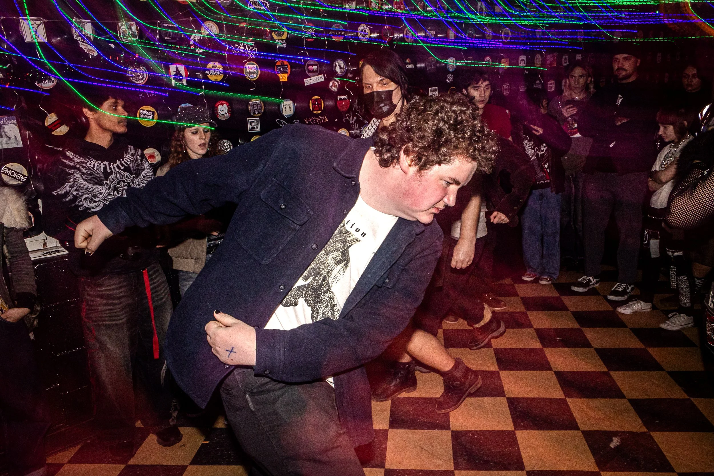 People dancing or participating in a party or nightclub scene with colorful neon string lights overhead and various stickers or patches on the background wall.