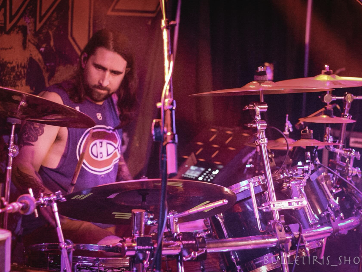 A drummer playing a drum set on stage with purple and pink lighting, wearing a sports jersey with an 'HC' logo, with drumsticks in hand and long hair.