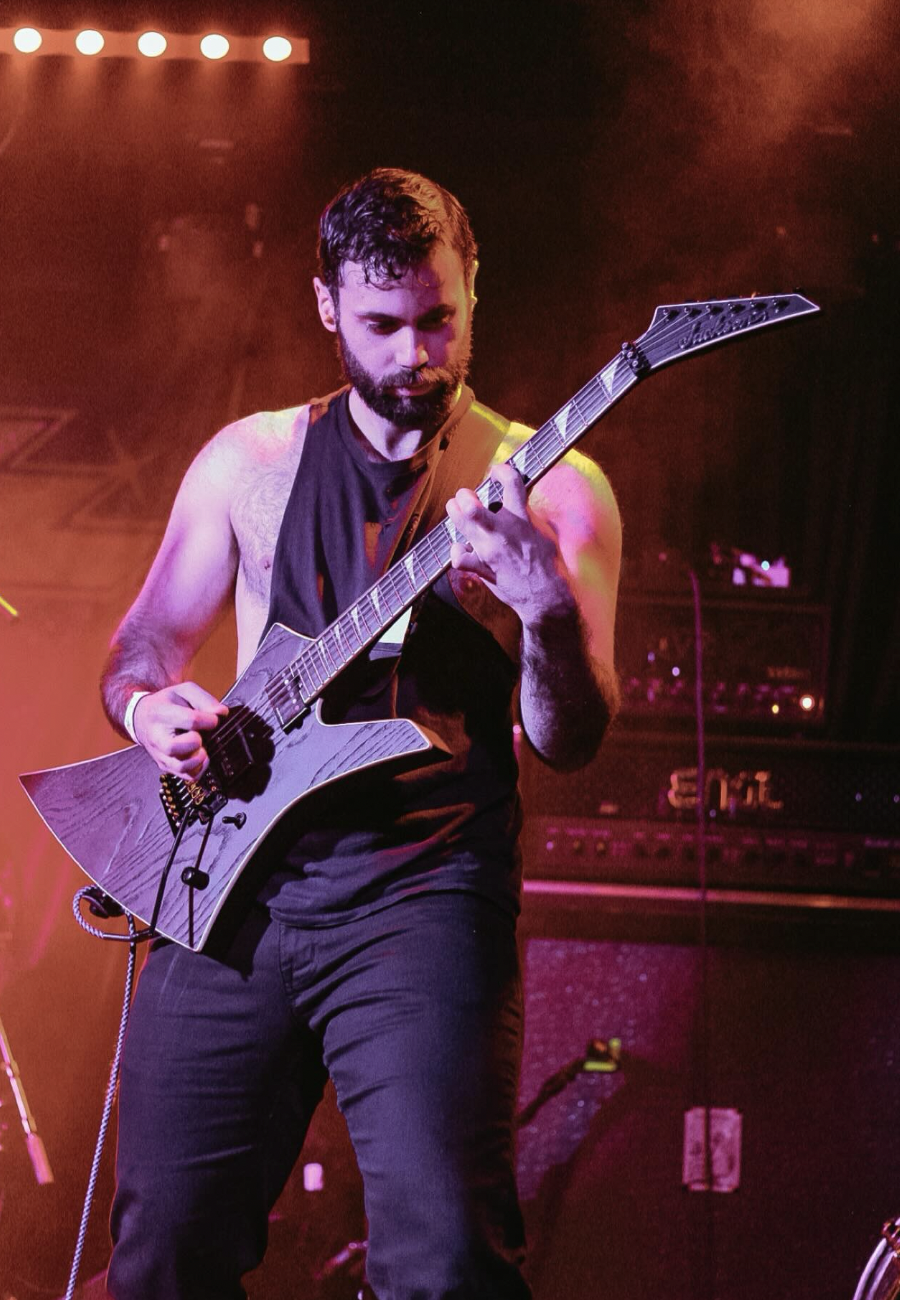A man with a beard and dark hair playing an electric guitar on stage with purple and orange lighting.