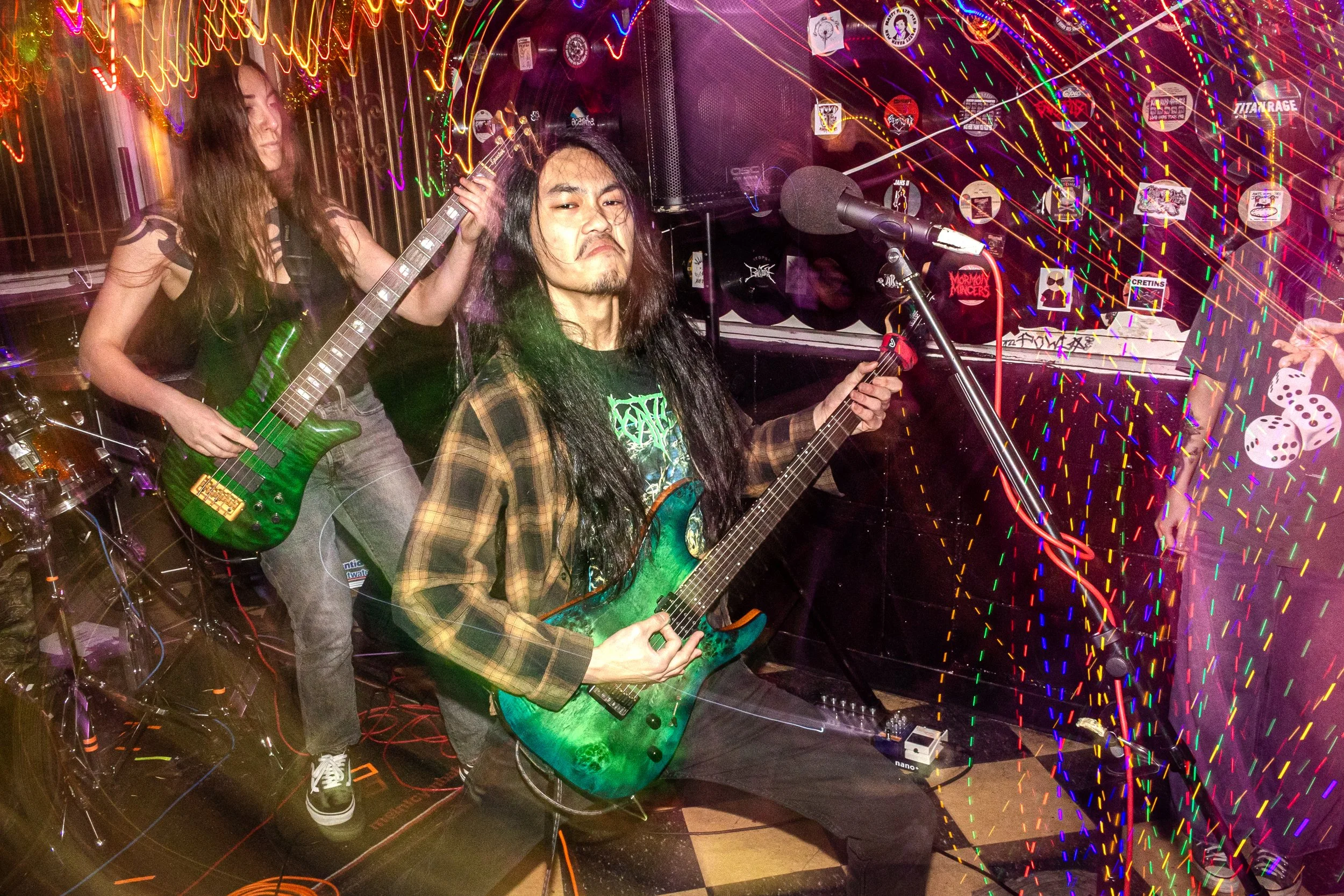 A band performing with two guitarists and a drummer in a neon-lit setting with colorful streaks of light and stickers on the wall