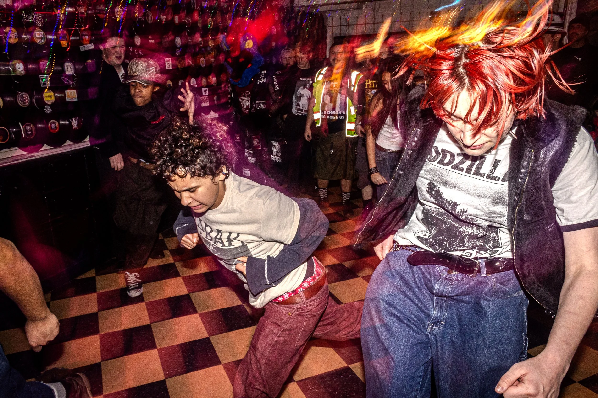People dancing and having fun inside a festive venue with colorful string lights and punk rock decor. One person with bright red hair is in motion, others are dancing or watching. The floor is a checkerboard pattern.