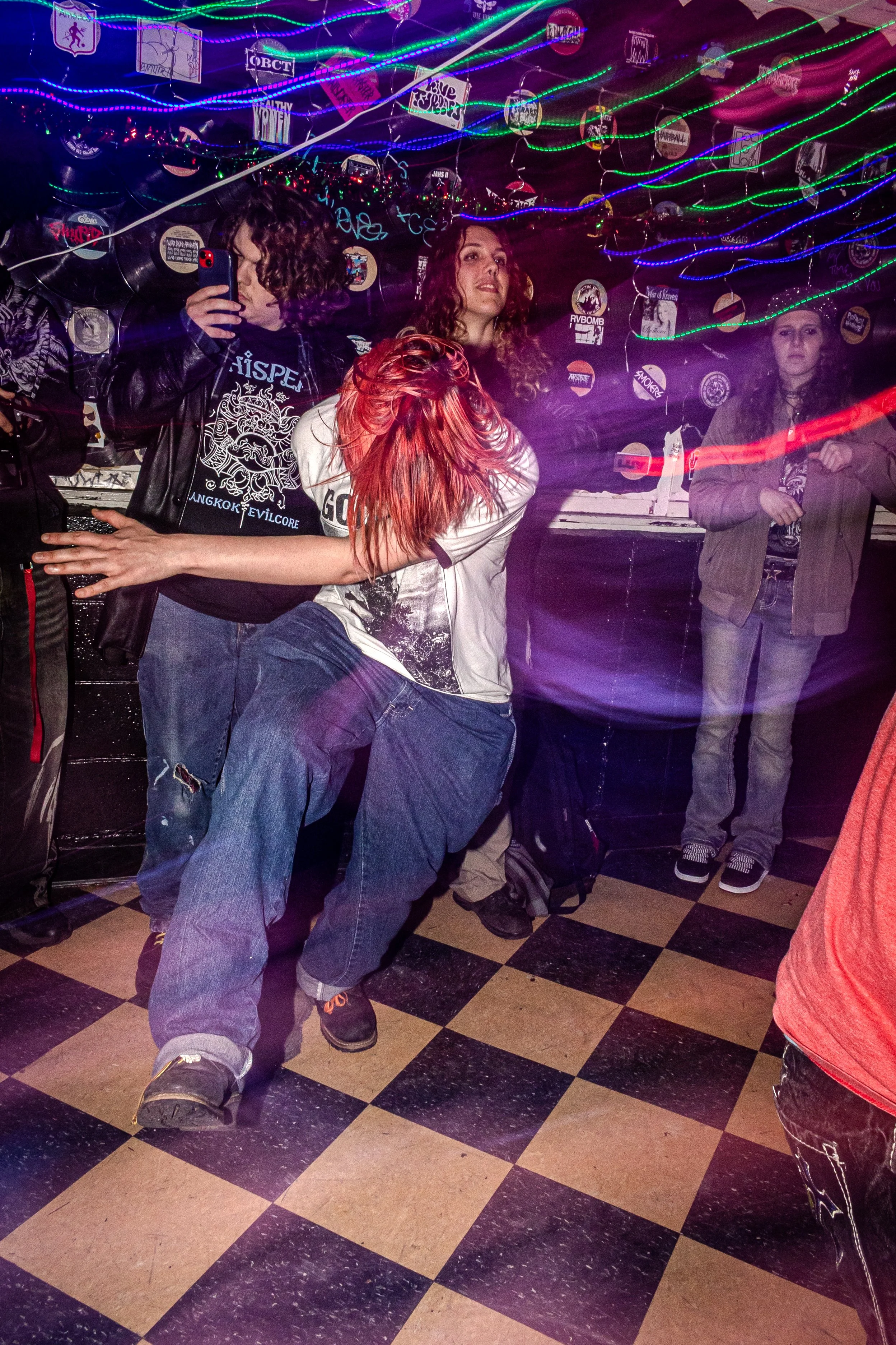 A group of people in a dimly lit nightclub or party setting with neon lights, posters, and vinyl records in the background. One person is taking a selfie, while others are dancing or standing around.