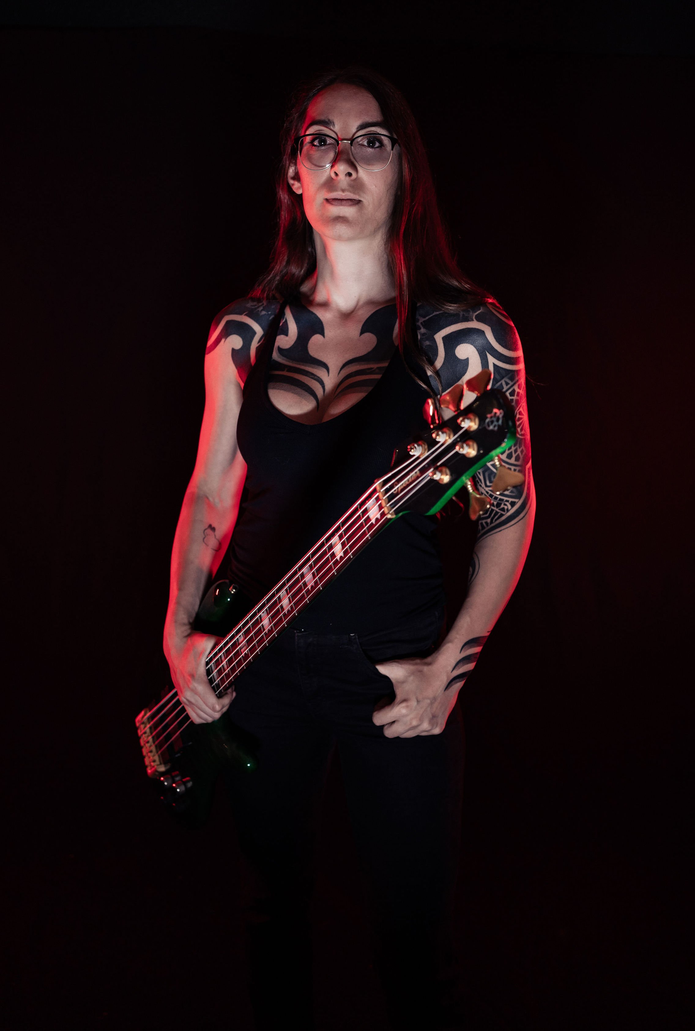A woman with long dark hair and glasses holding a guitar. She has tattoos on her arms and chest, and is wearing a black top against a dark background with red lighting.