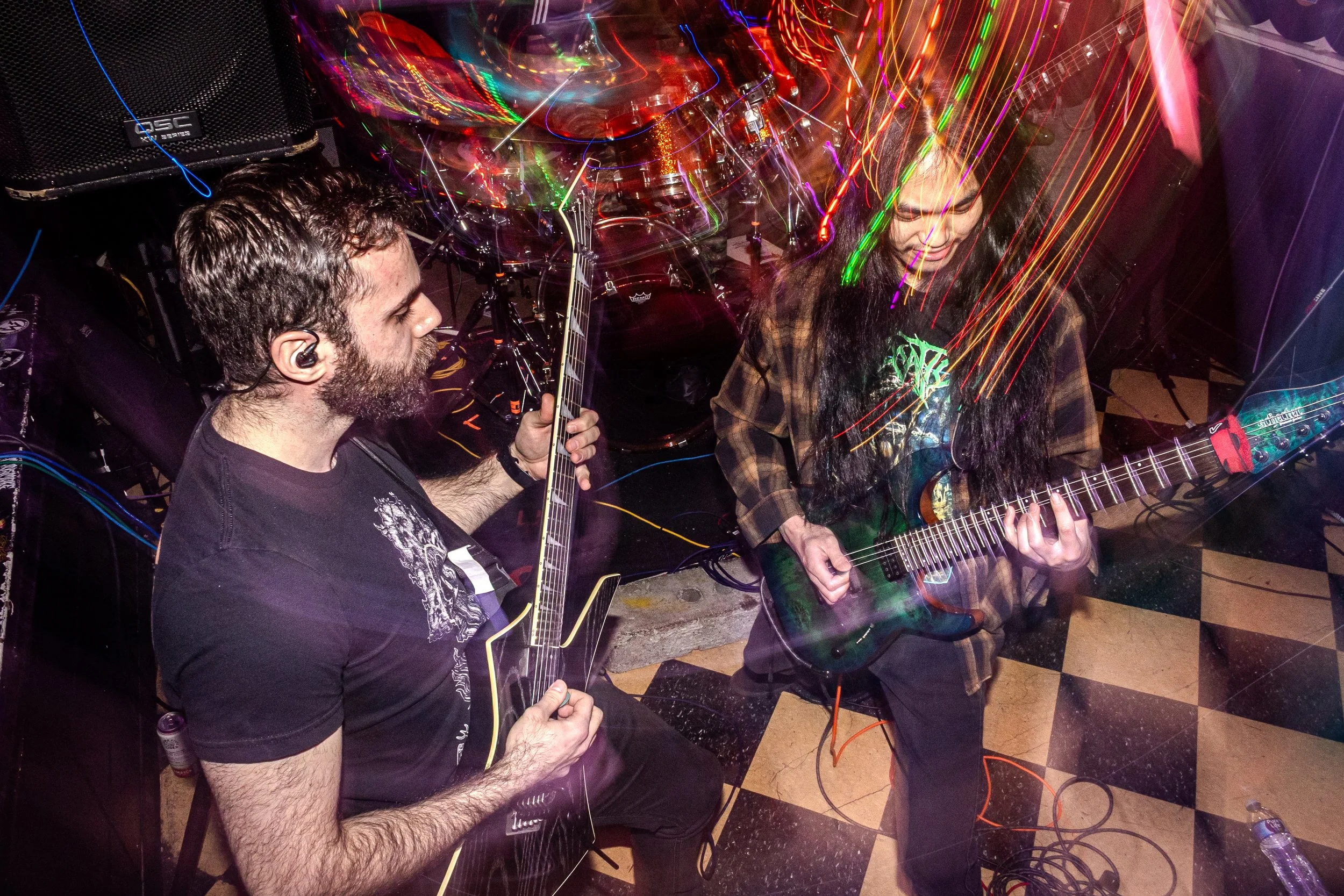 Two musicians playing guitars at a live performance with colorful light trails and a speaker in the background.