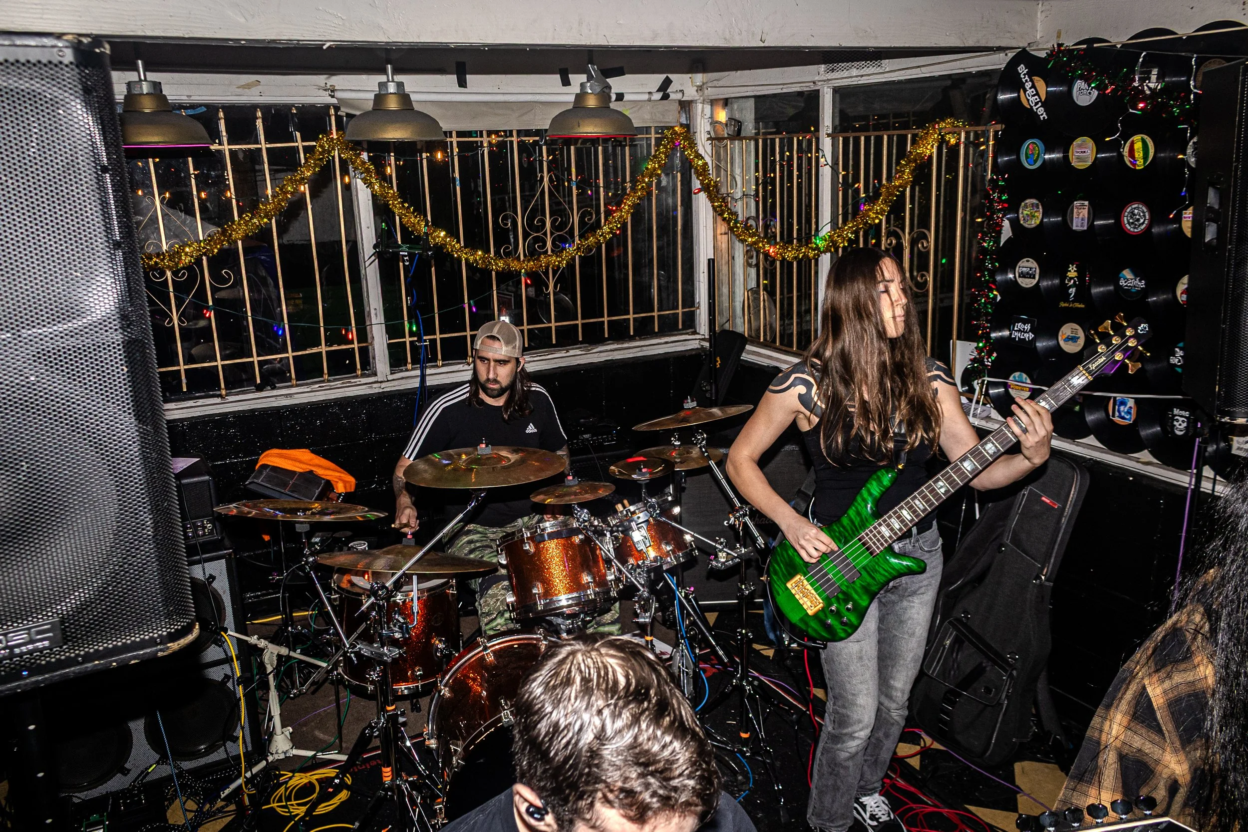 Band performing live in a decorated venue, with Christmas lights and tinsel, featuring a drummer and a bassist playing guitar.