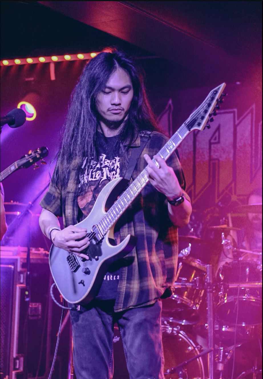 A male guitarist with long black hair playing an electric guitar on stage with purple and pink lighting, and a drum set in the background.