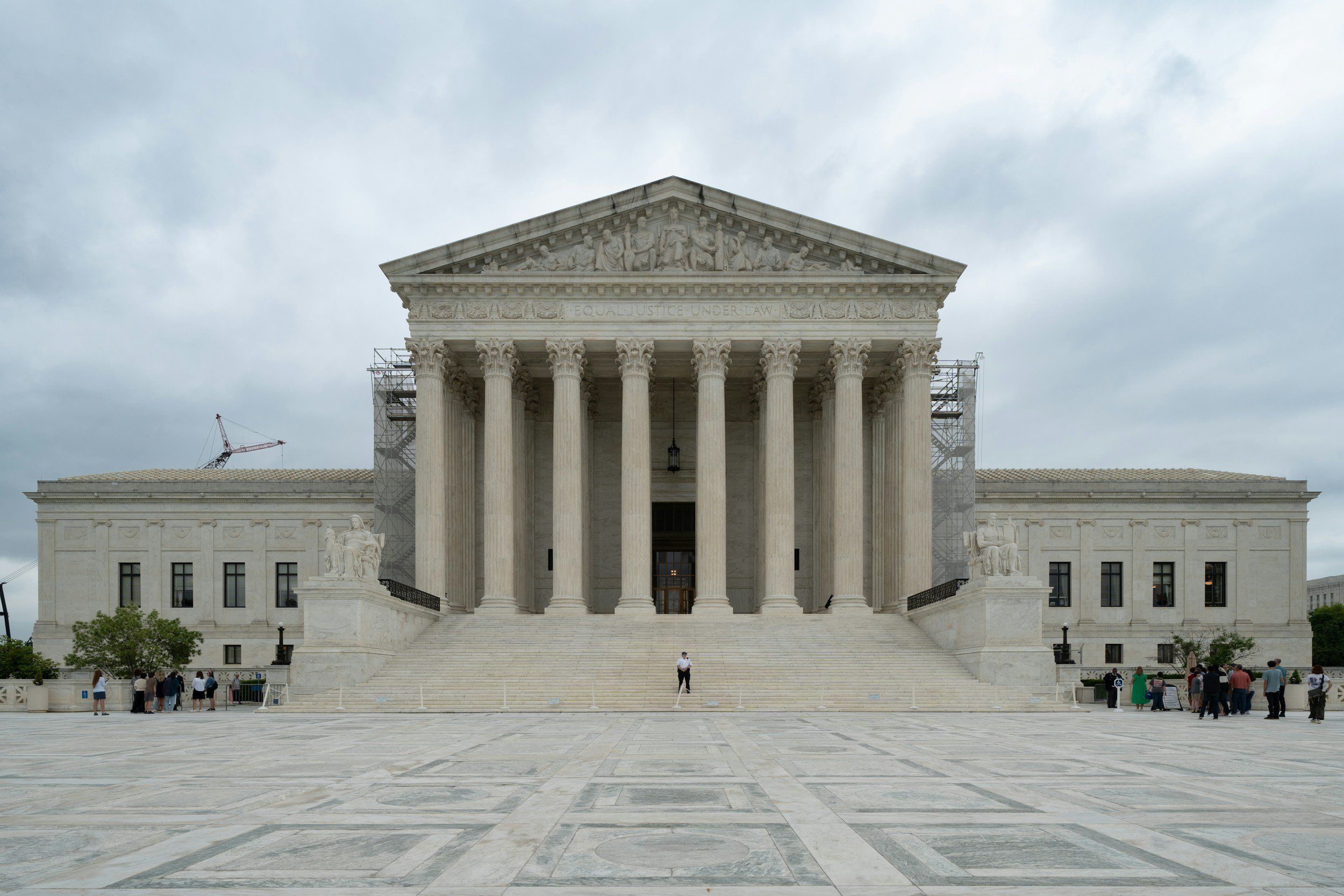 The U.S. Supreme Court building with neoclassical architecture, large columns, and statues on either side of the stairs, under a cloudy sky, with people walking in front.